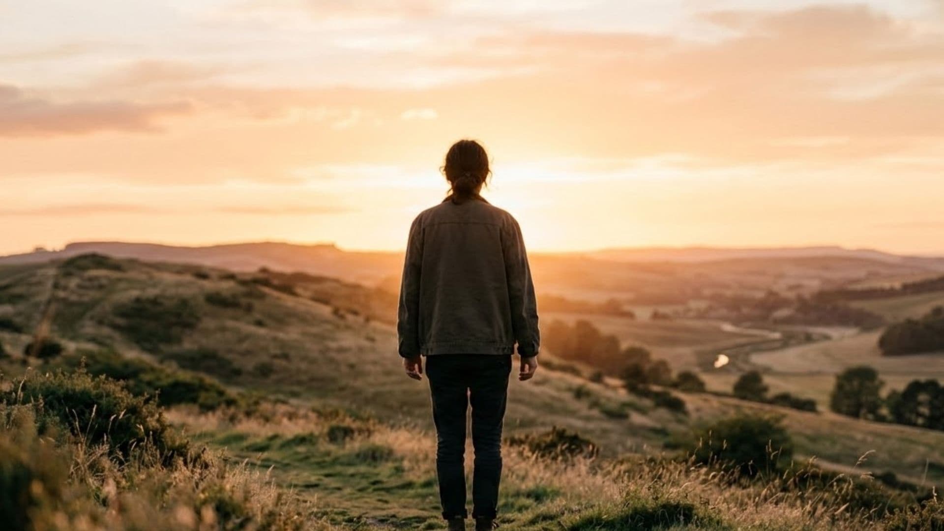 a person in a brown jacket and dark jeans standing on a grassy hilltop facing a wide valley at sunset