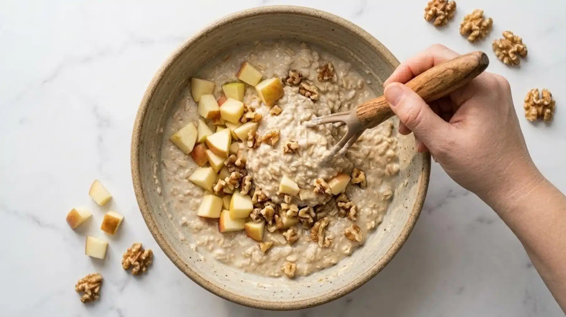 a person stirring a bowl of oatmeal topped with apple chunks and walnuts using a wooden fork on a marble surface