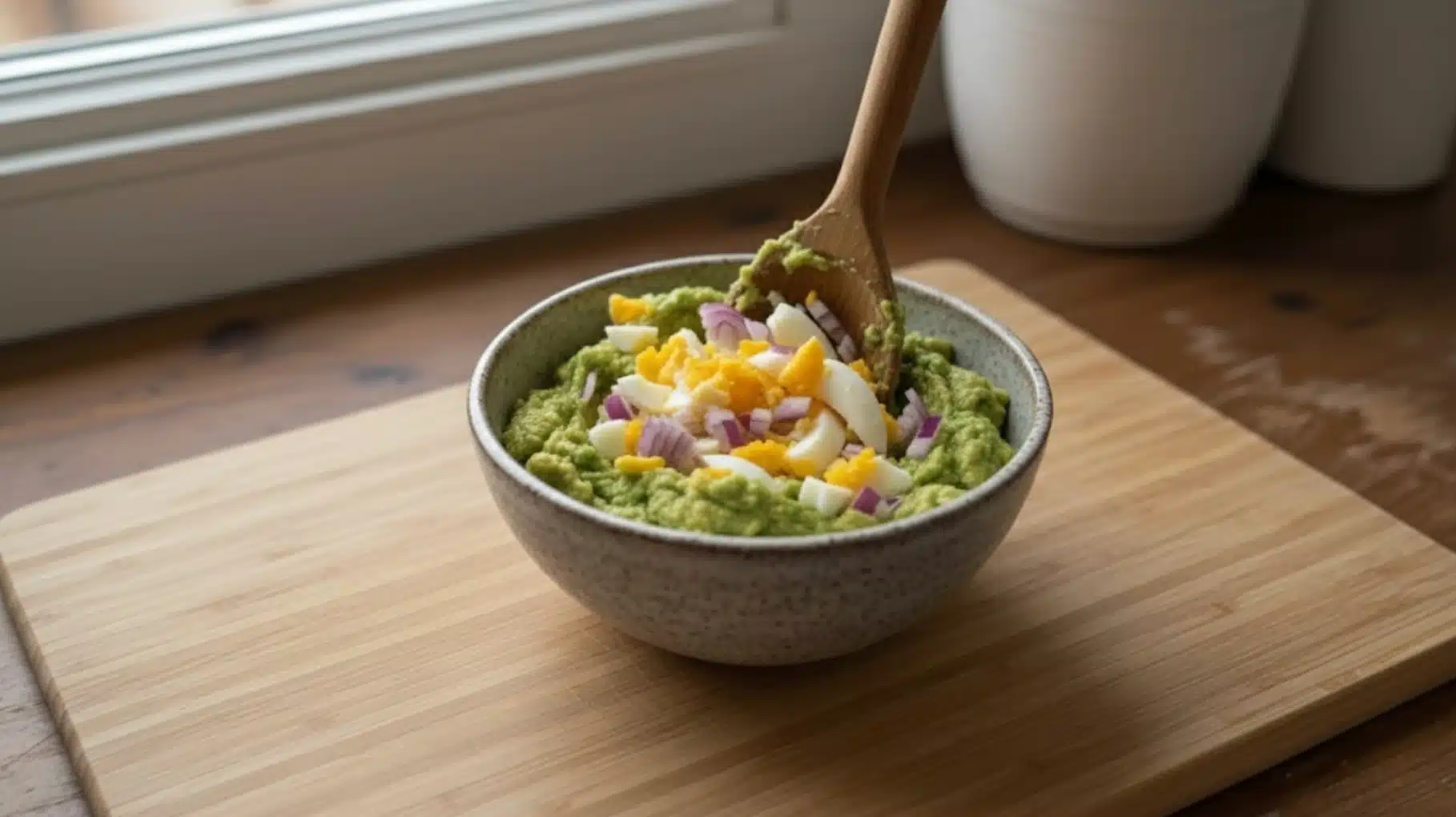 bowl of guacamole topped with chopped boiled eggs and red onion, stirred with a wooden spoon, placed on a wooden surface