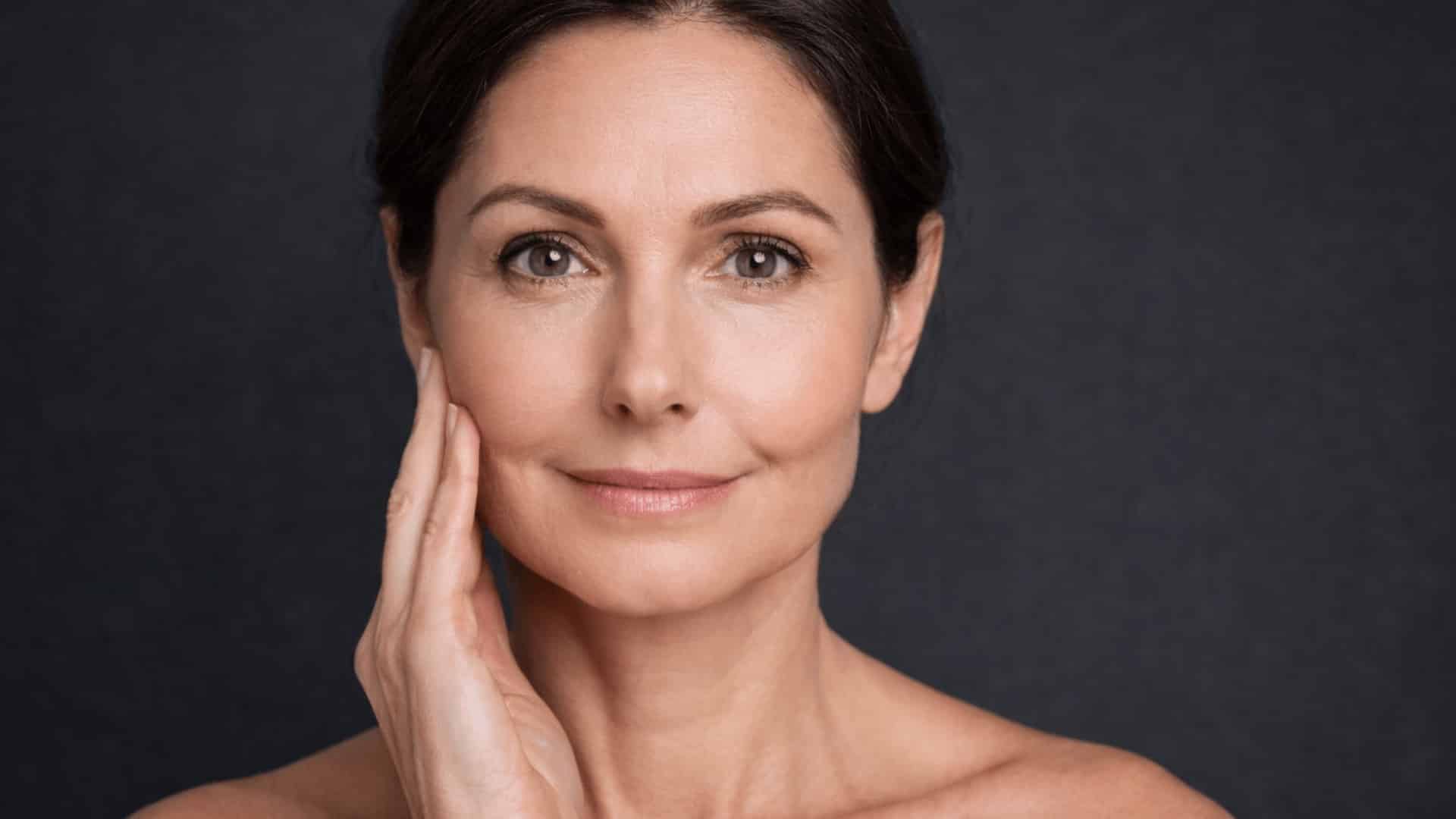 brunette middle aged woman gently touching her cheek against charcoal grey background with soft lighting