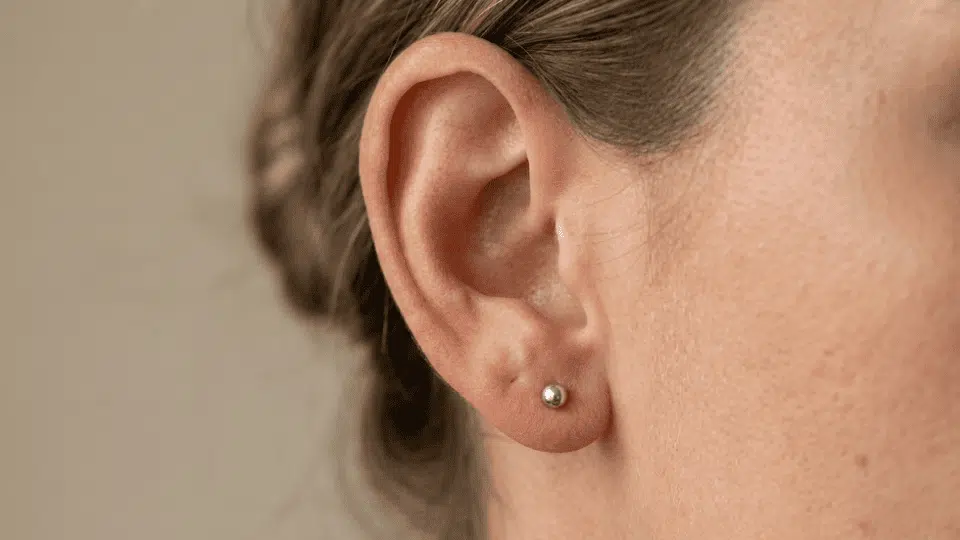 close-up of a woman’s ear with a simple silver stud in the lower earlobe against a soft neutral background