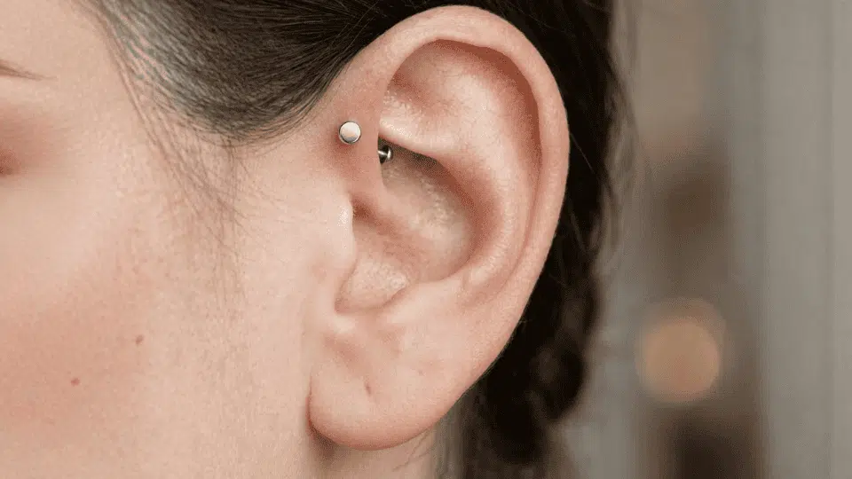 close-up of an ear featuring a silver forward helix piercing on the front upper cartilage, hair pulled back, soft indoor background