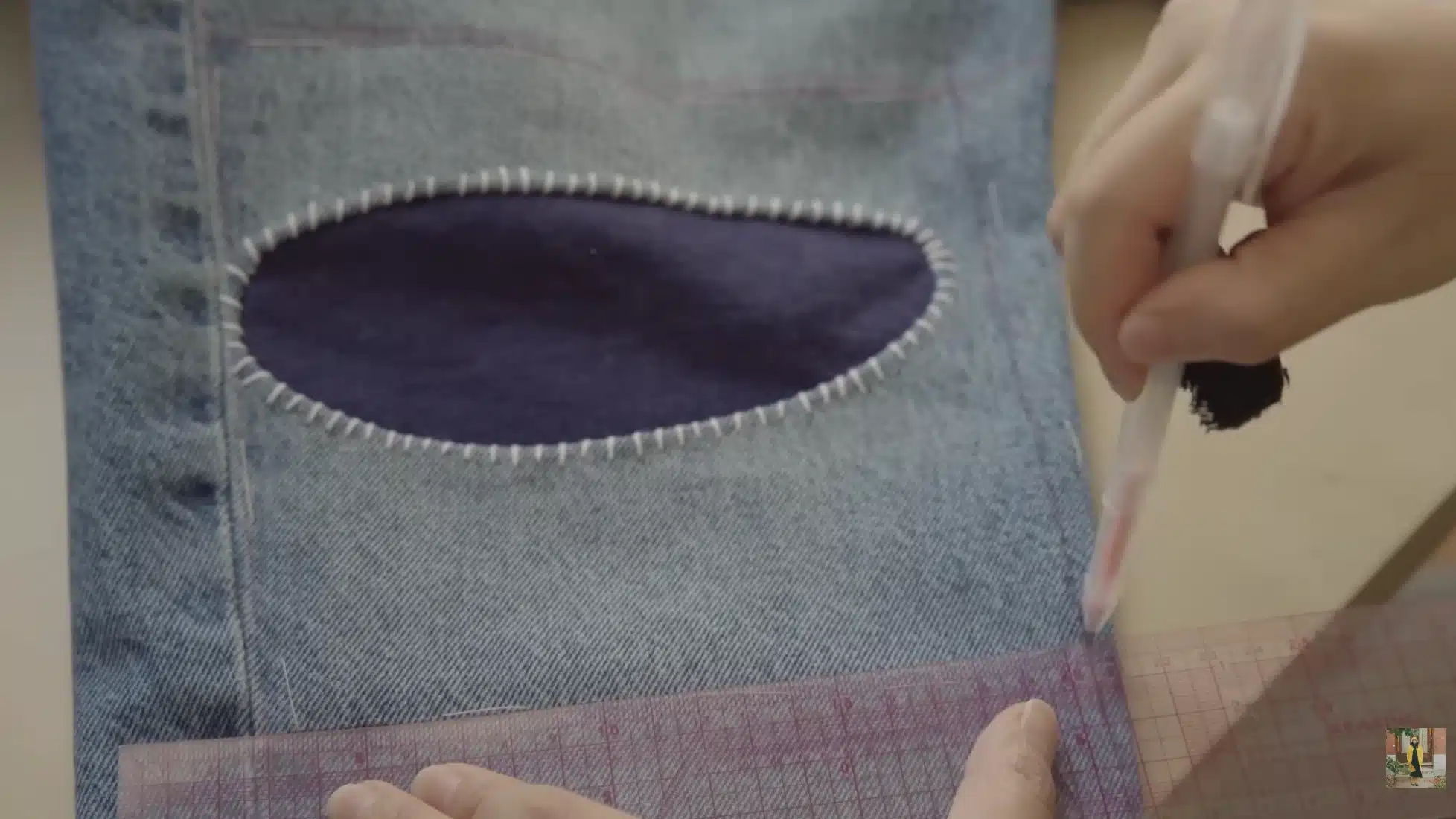 denim jeans with oval patch stitched around hole while hands mark sashiko stitching grid using ruler and fabric marker