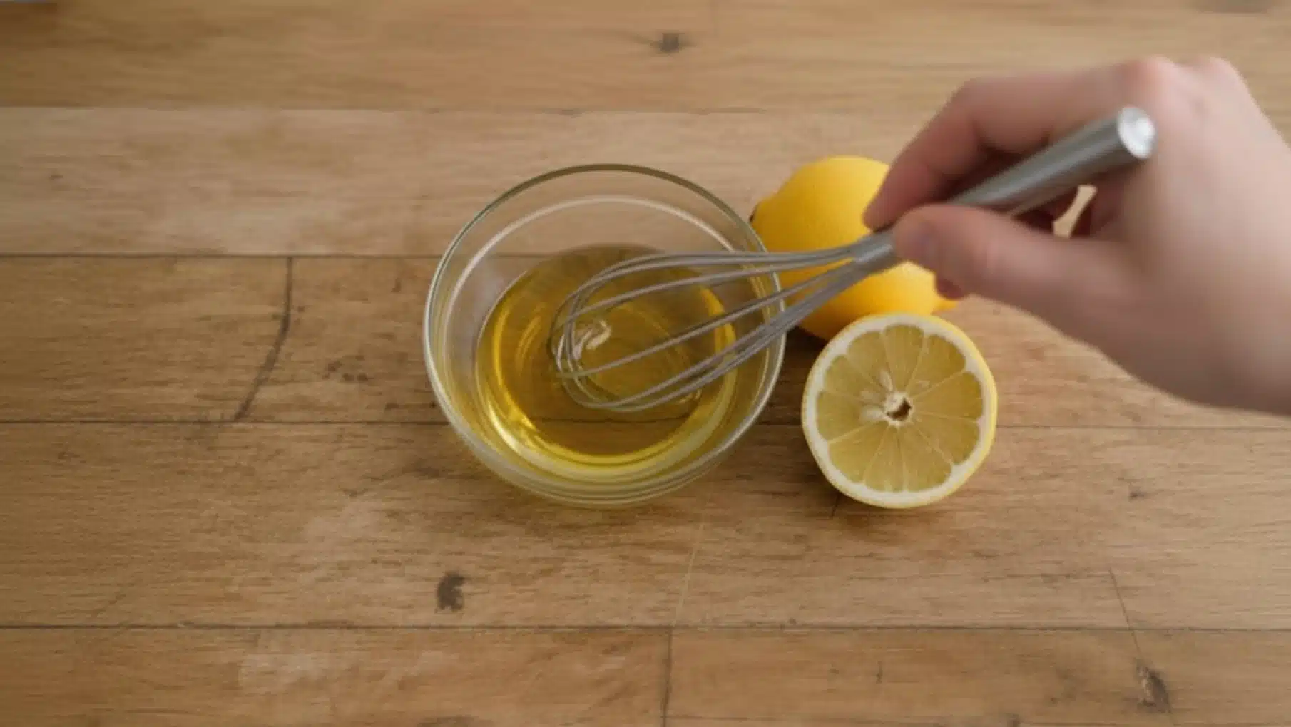 hand whisking olive oil and lemon juice in small glass bowl on wooden table, with whole lemon and half beside