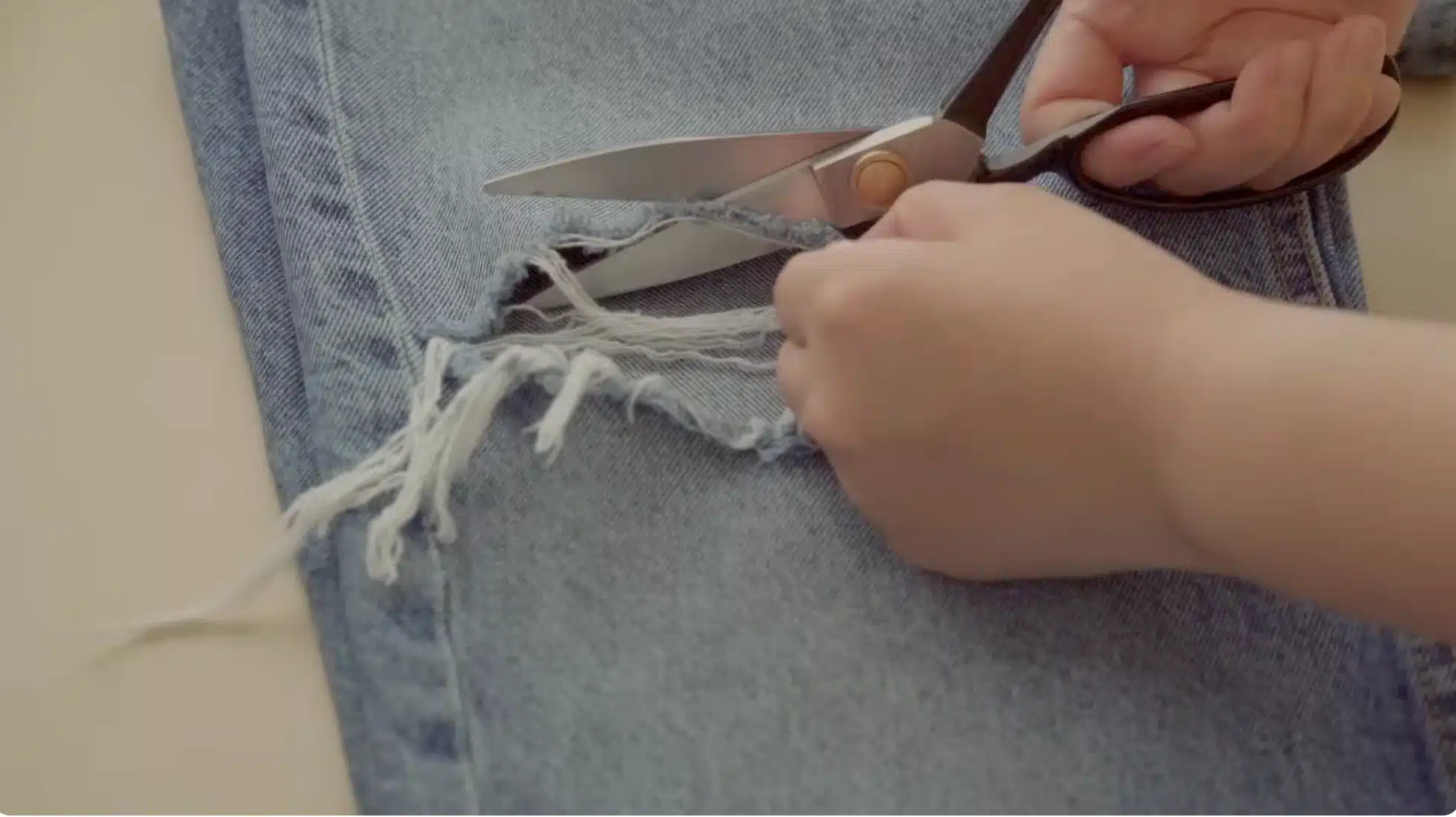 hands cutting frayed threads from torn denim jeans using scissors, preparing damaged fabric area for patch repair