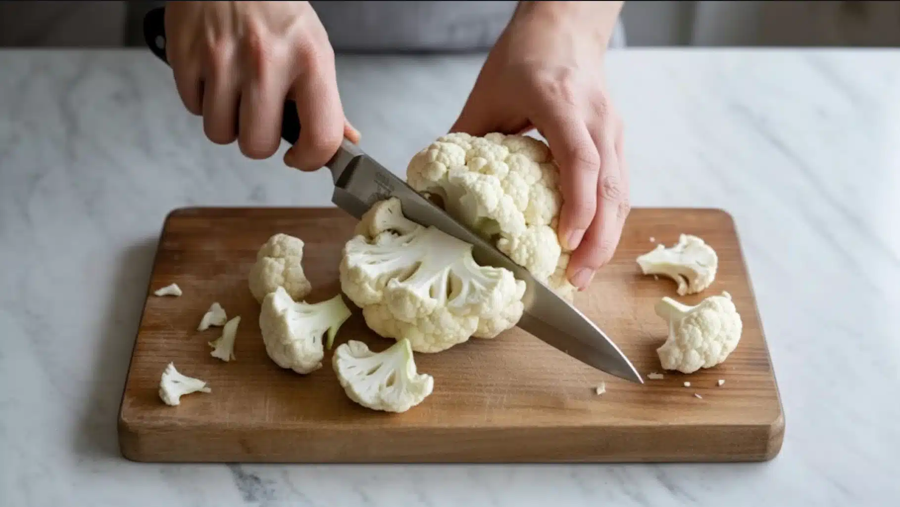 hands cutting fresh cauliflower into small florets on a wooden cutting board using a kitchen knife on marble countertop