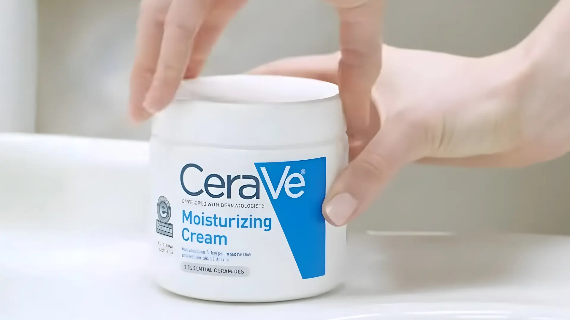 hands lifting the white cap off a cerave jar to remove the lid of the cream sitting on a bathroom sink