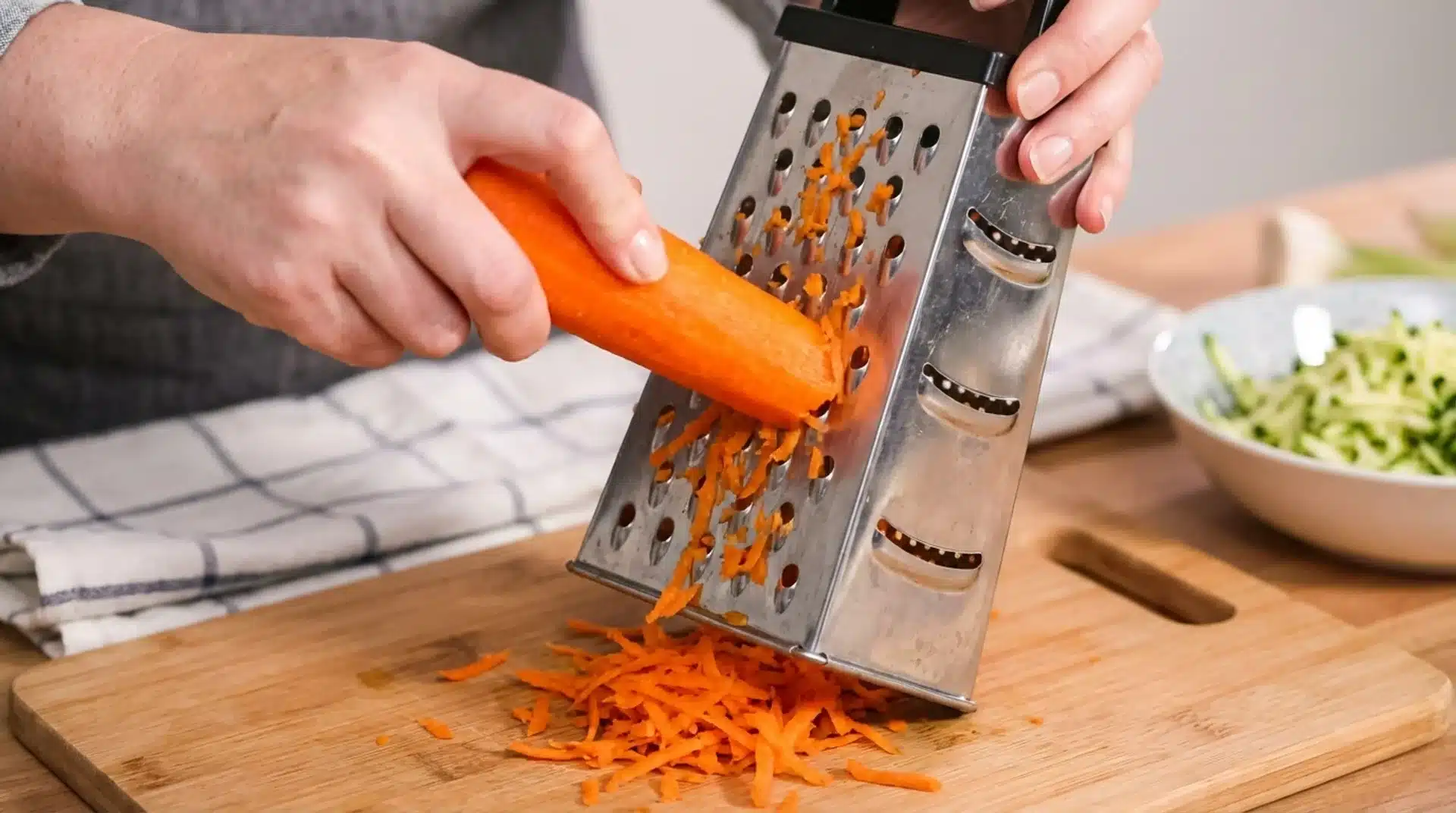 person grating fresh carrot on metal box grater over cutting board, with shredded carrot pieces and vegetables nearby