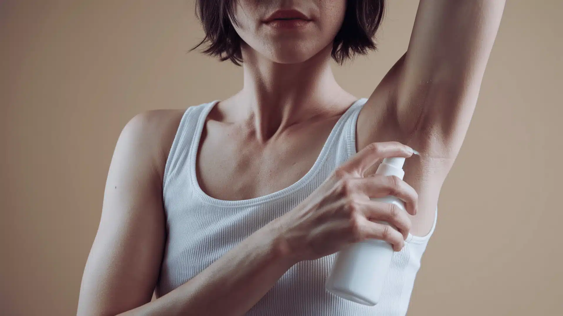 person with short and no face in the frame, wearing a white tank top she is holding a white spray bottle