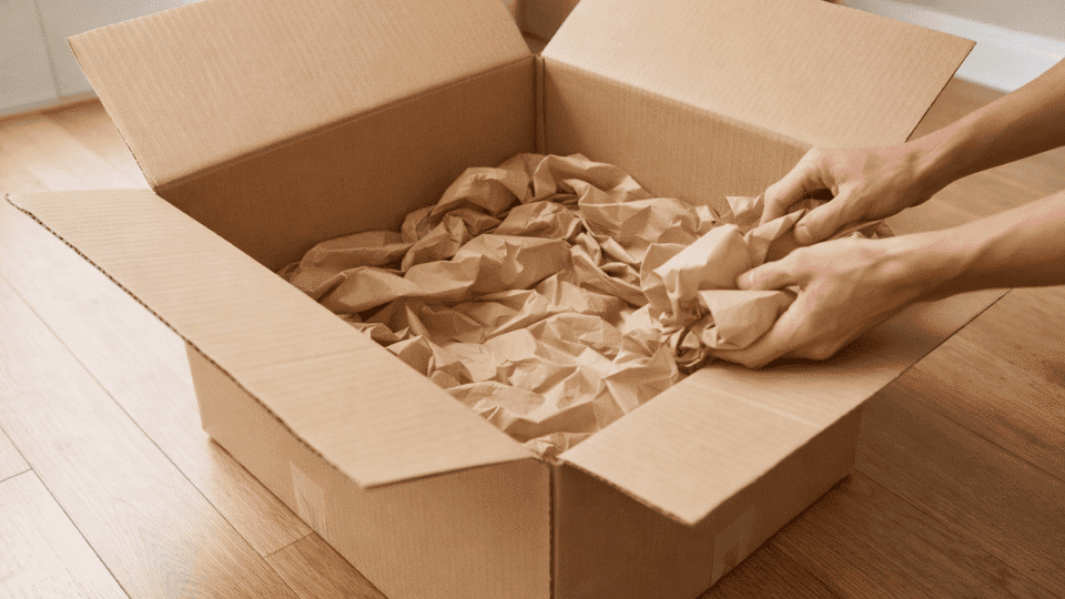 placing crumpled packing paper inside a taped cardboard moving box to cushion fragile items before packing glasses for a move
