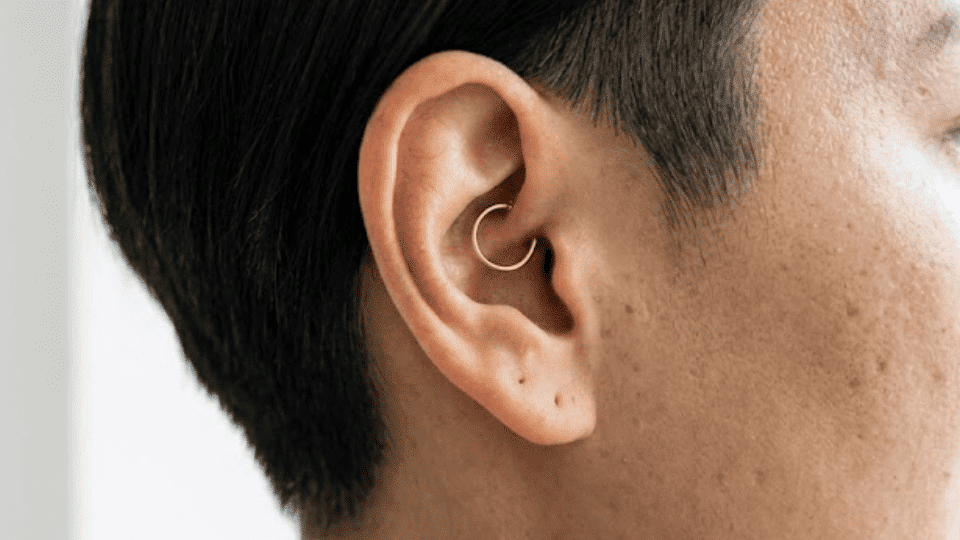 side profile of ear with a gold daith ring inside inner cartilage, short hair, bright window light background