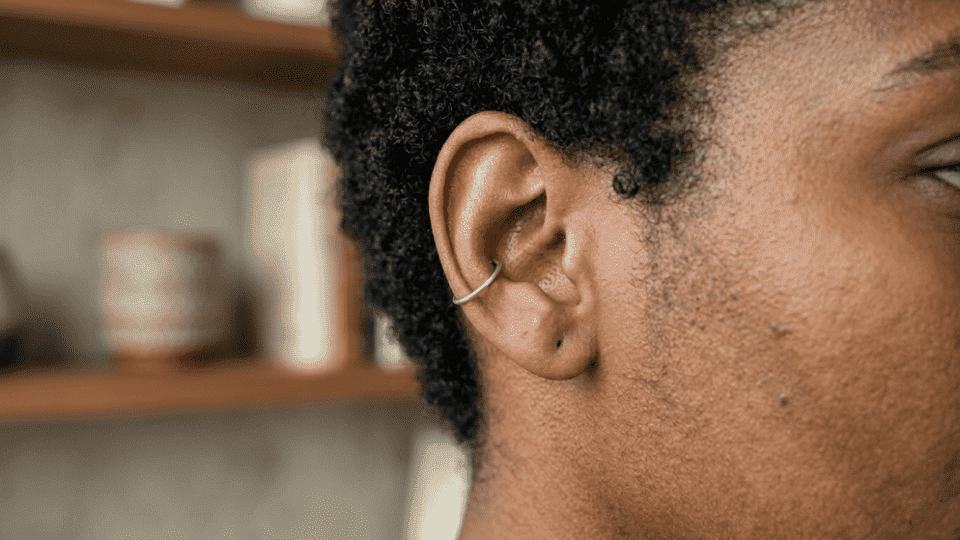side profile of ear with a silver outer conch hoop, short curly hair, soft indoor light and blurred shelf background