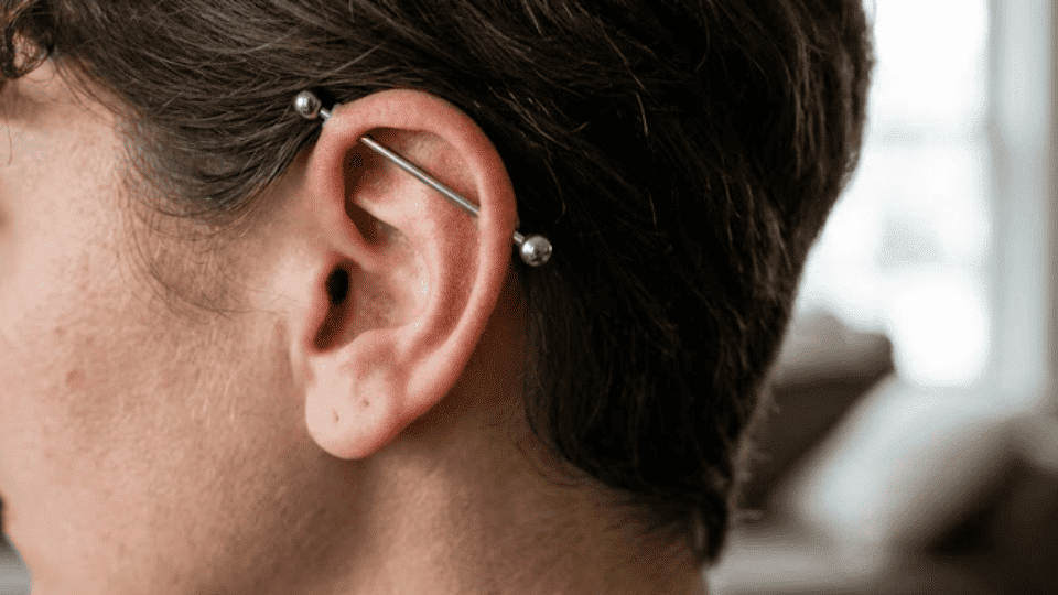 side profile of ear with industrial bar across upper cartilage, short hair, bright living room background