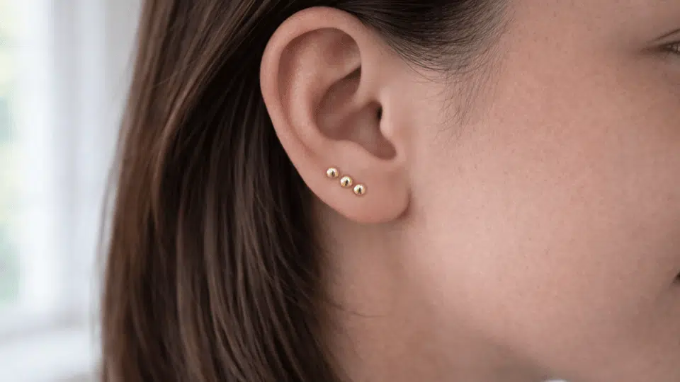 side profile of ear with three small gold stacked lobe studs, hair tucked back, soft indoor daylight background