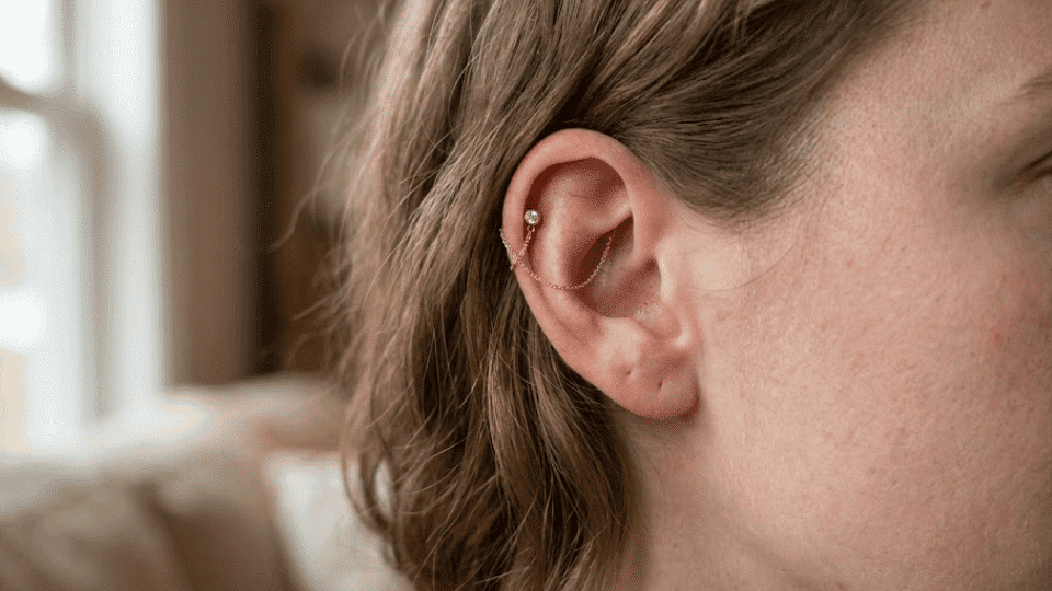 side profile of woman with hidden helix gold stud and chain, soft window light and cozy indoor background