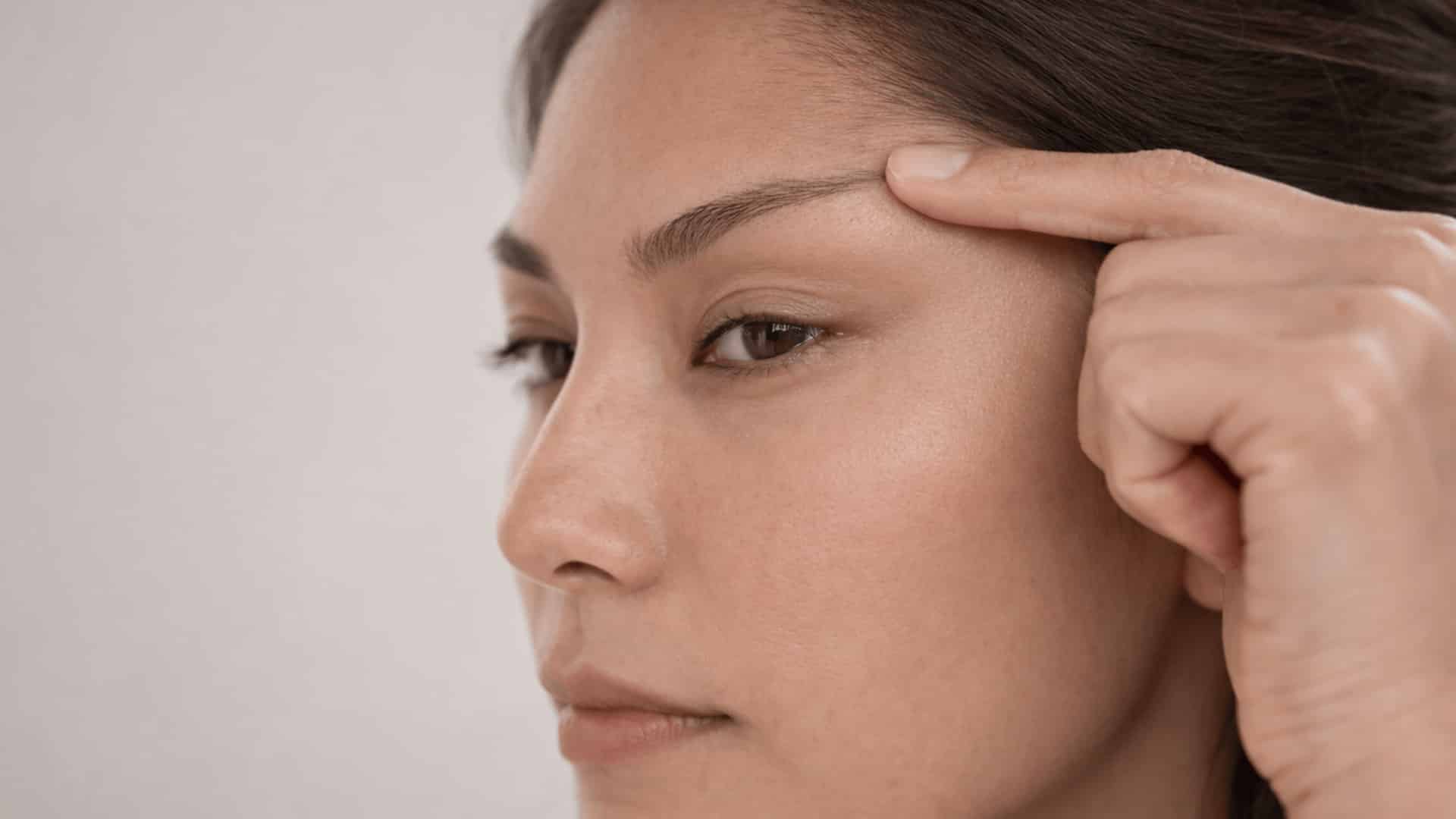 three quarter side profile of woman pressing brow bone to check depth of hooded eyes under soft studio lighting