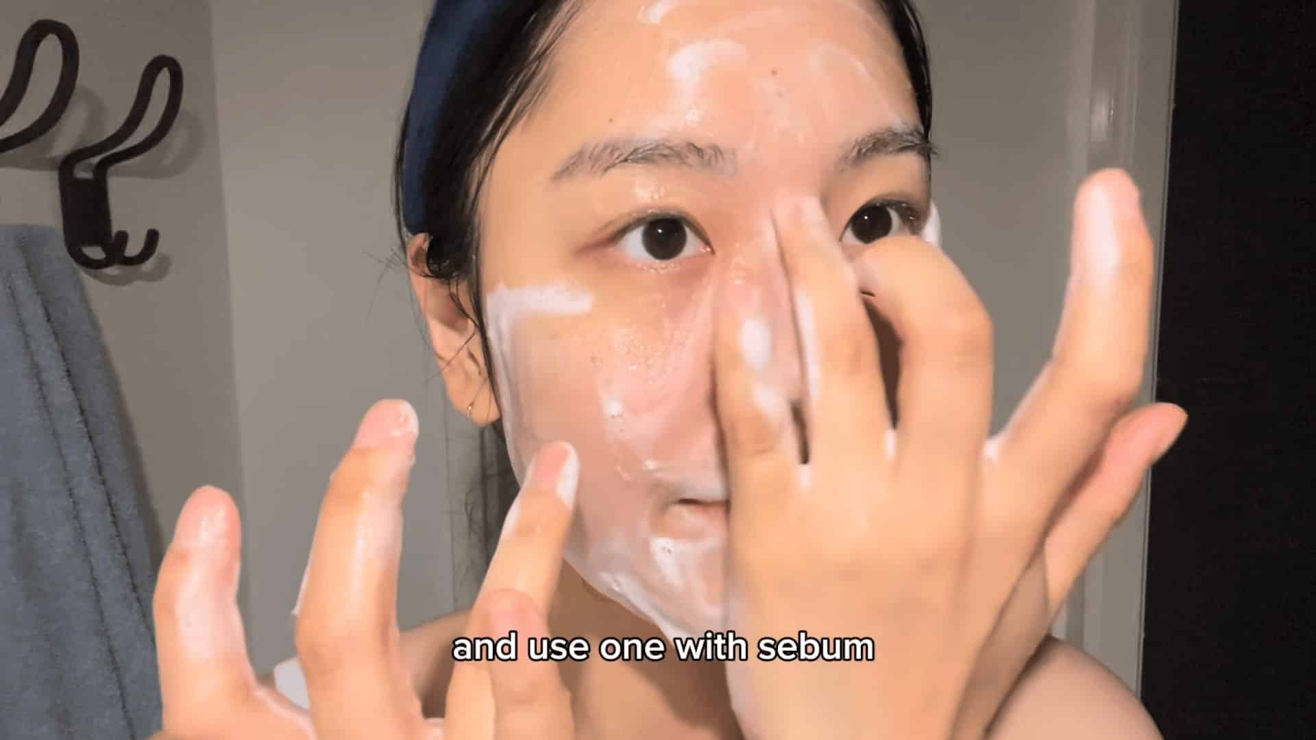 woman applying foaming cleanser to face in bathroom, massaging cheeks and forehead with both hands