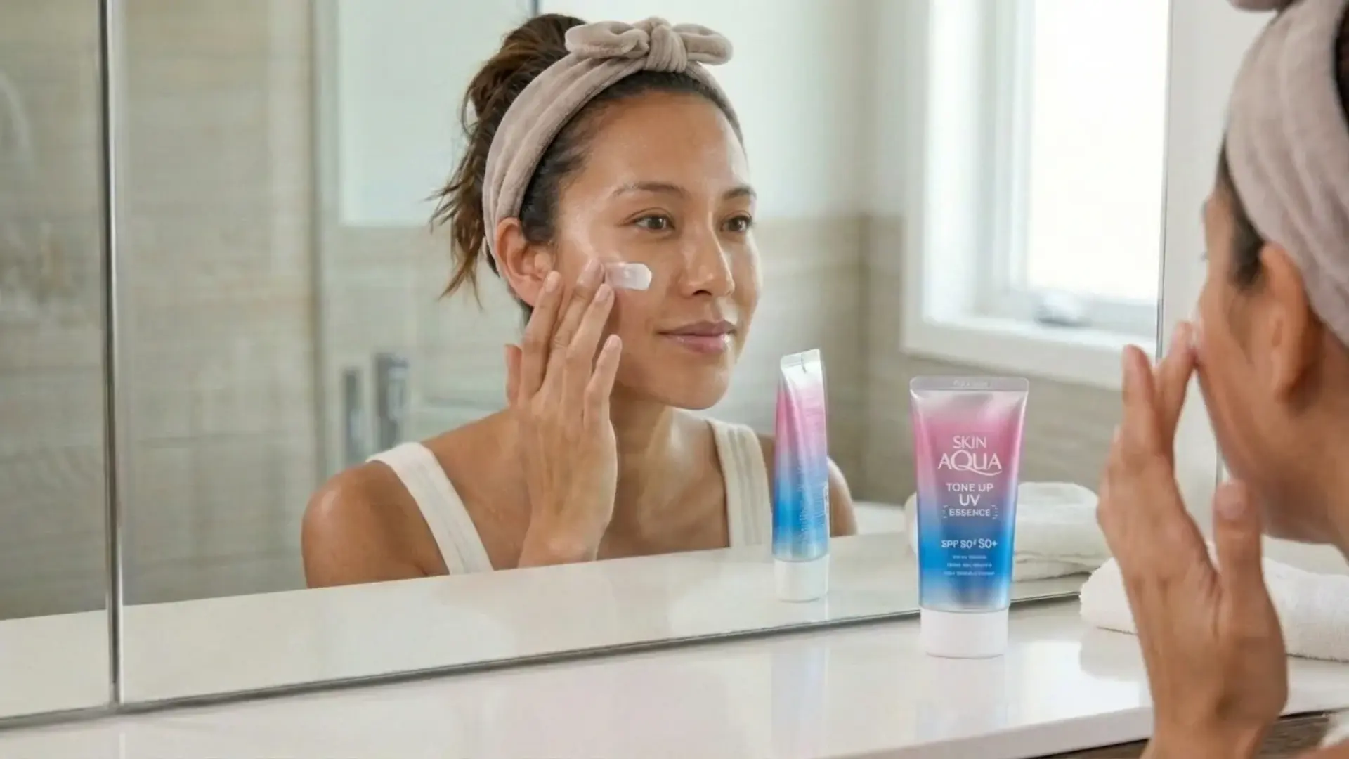 woman applying sunscreen (am only) to her face in a mirror with pink and blue gradient tubes on the counter