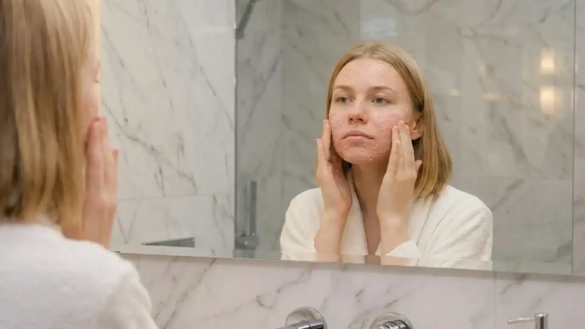 woman in a white bathrobe massaging a gentle cleanser into her dry skin in a bright bathroom