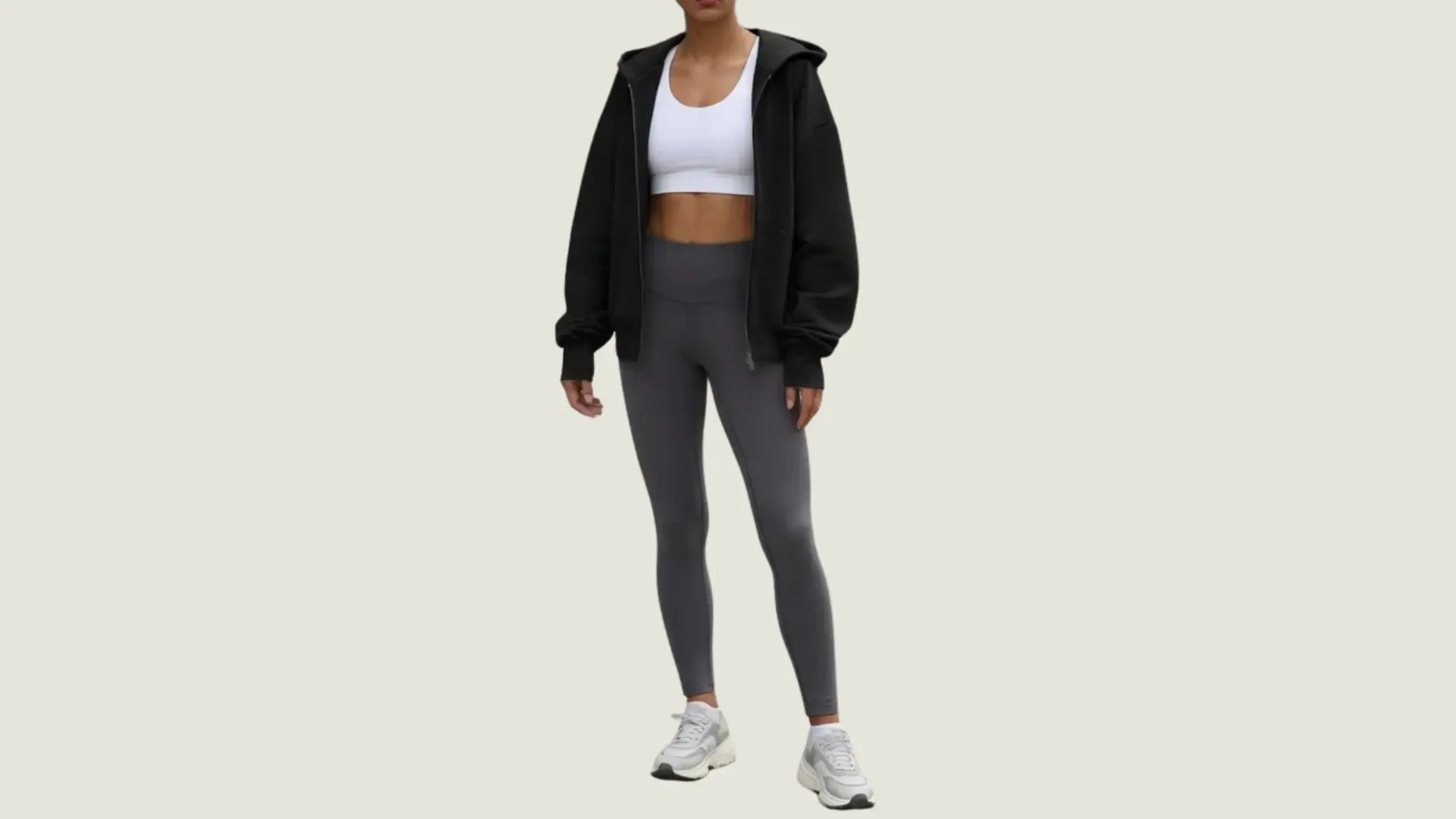 woman in an athleisure style wearing a white sports bra, black zip-up hoodie, grey leggings, and white sneakers