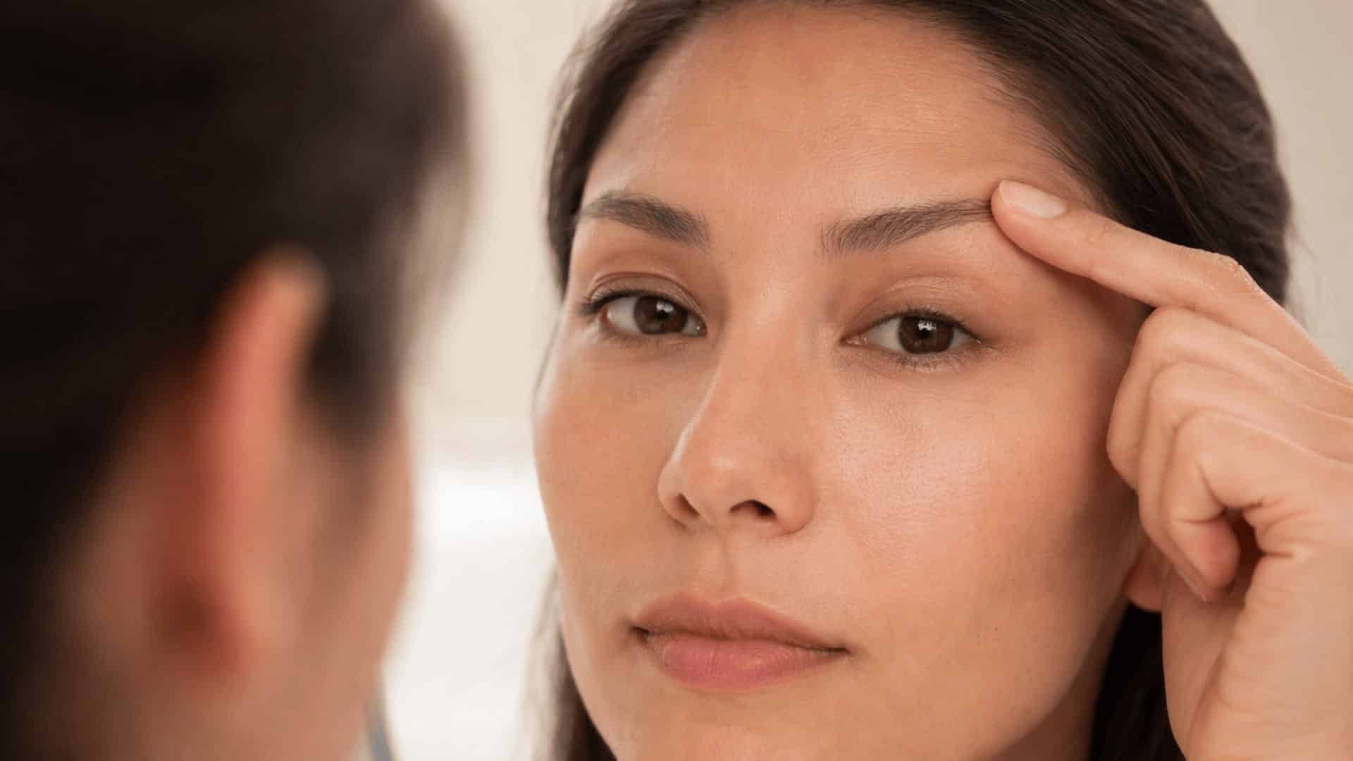 woman lifting brow in mirror to reveal hooded eyelid crease under soft bathroom lighting