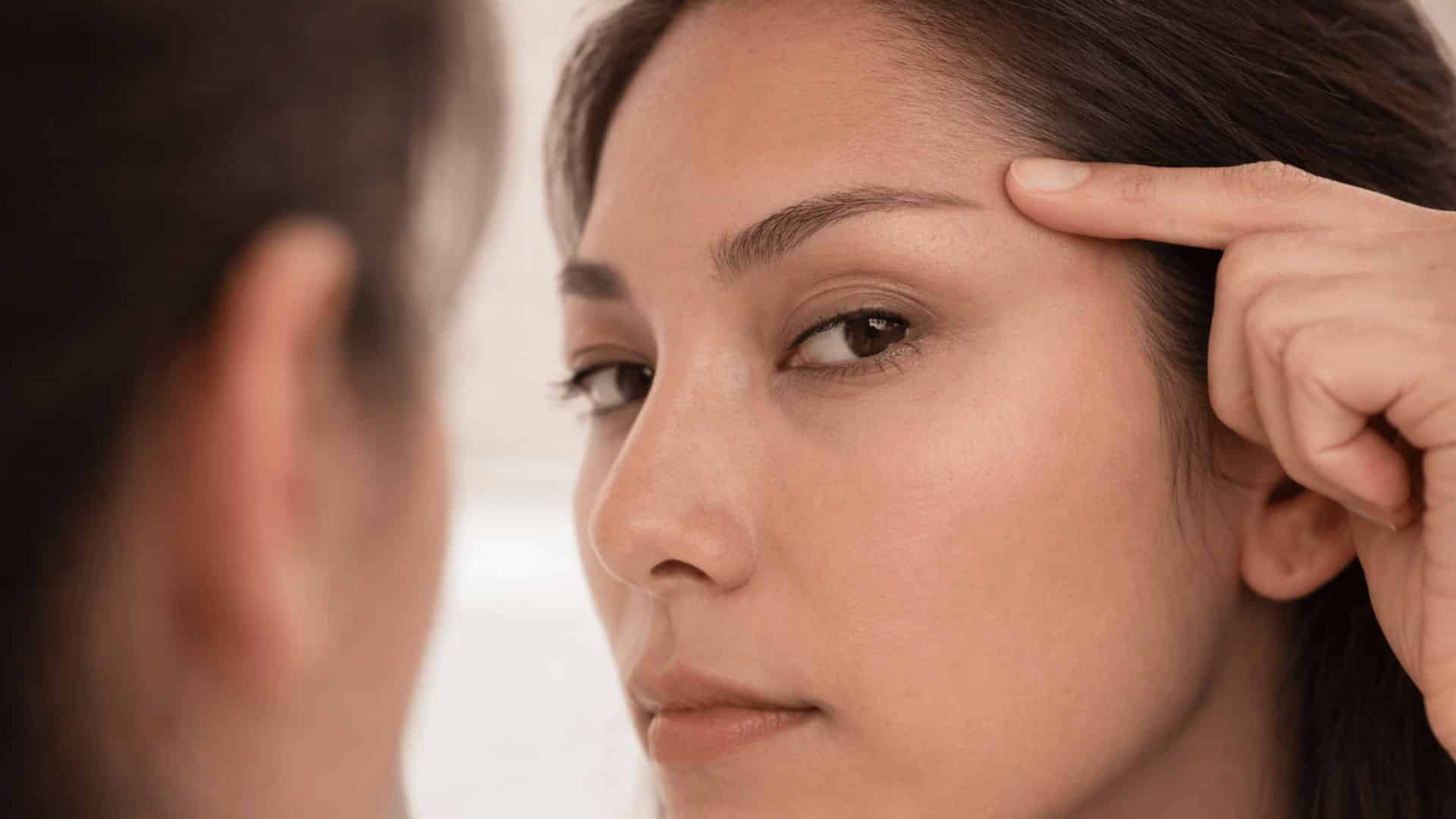 woman looking at outer corner of her eye in mirror under soft bathroom lighting with neutral expression