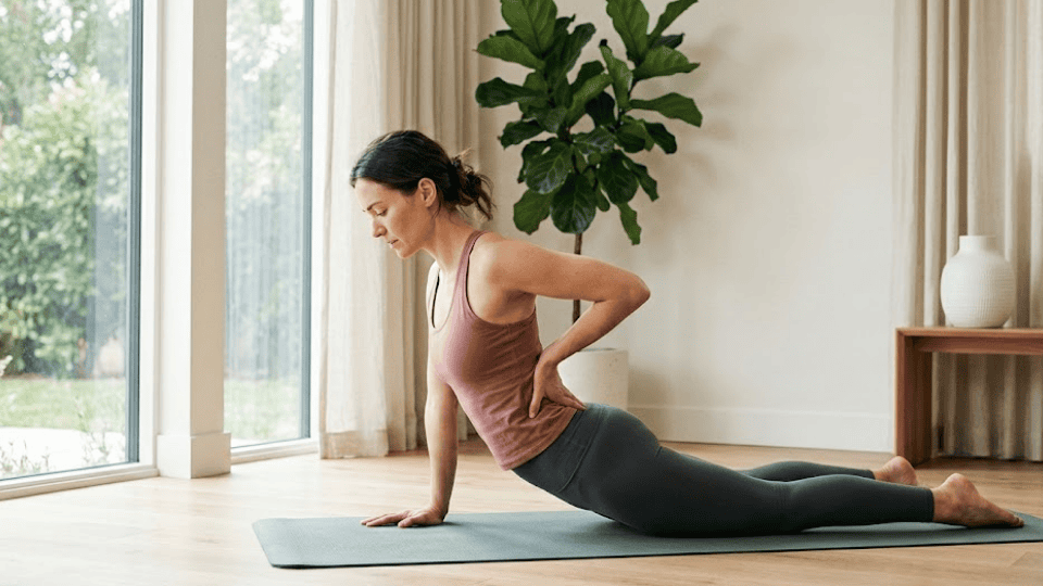 woman practicing yoga on a mat indoors, touching her lower back with a strained posture in a bright, calm living room setting