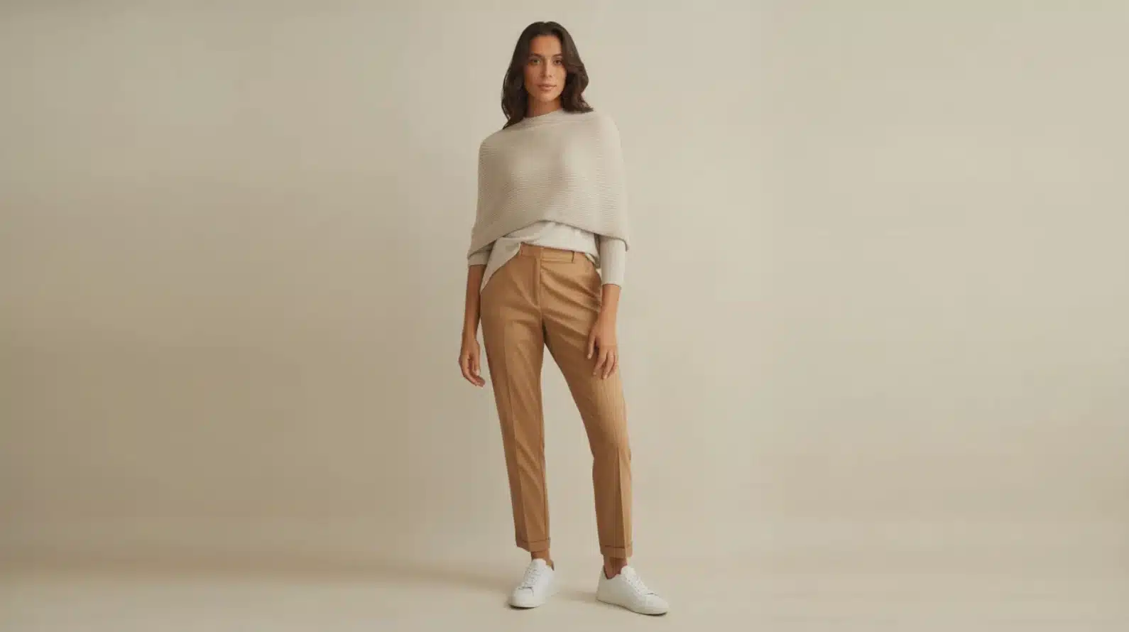 woman wearing beige knit top, tan tailored trousers, and white sneakers against neutral studio background
