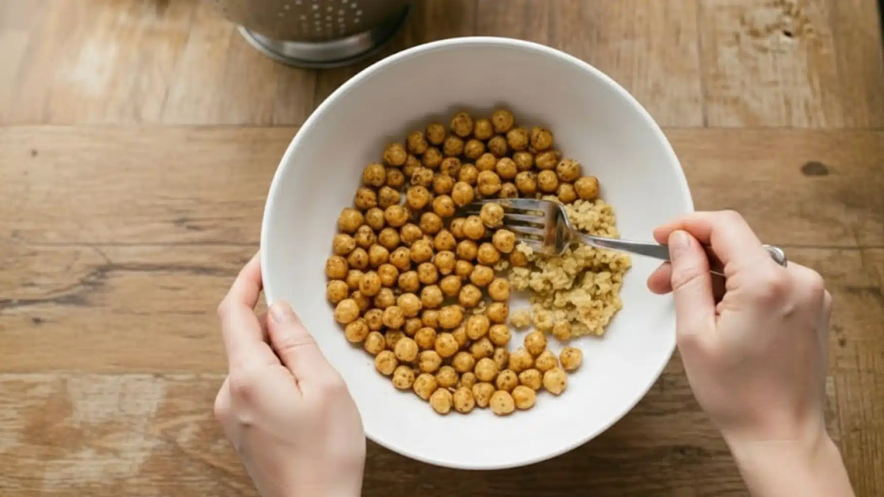 hands holding white bowl with roasted chickpeas and quinoa, using fork to mix food, placed on wooden table
