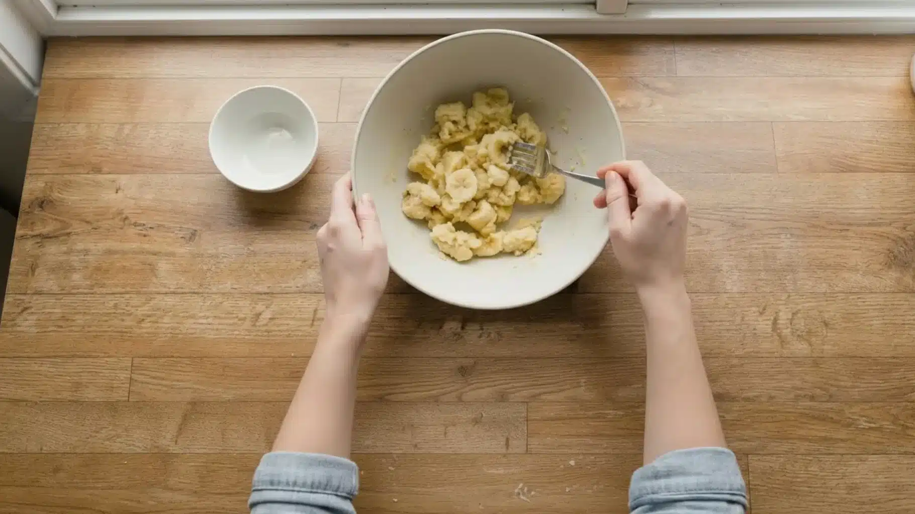 hands mashing ripe bananas with a fork in a mixing bowl on wooden countertop, preparing banana mixture for baking recipe