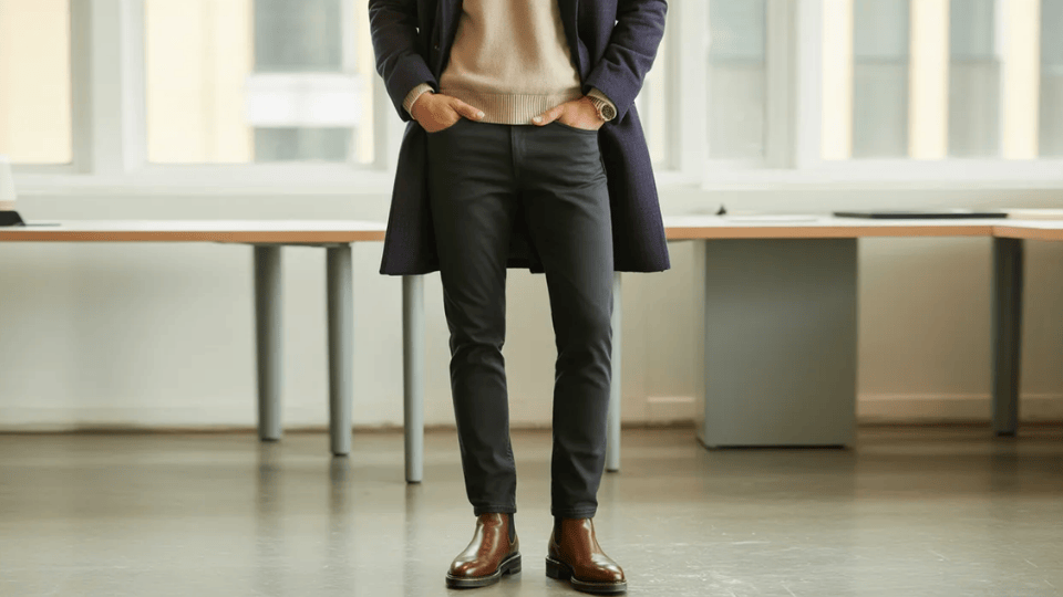 man wearing black jeans, knit sweater, and long coat with brown Chelsea boots in a modern office, showing a smart casual business outfit