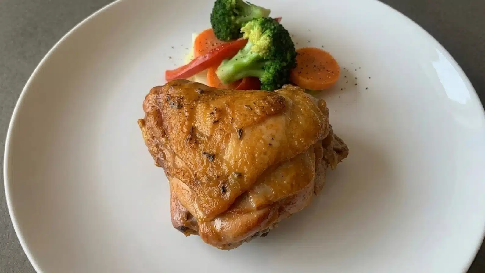 roasted chicken thigh with crispy skin served on white plate, side of broccoli, carrots, and red pepper slices