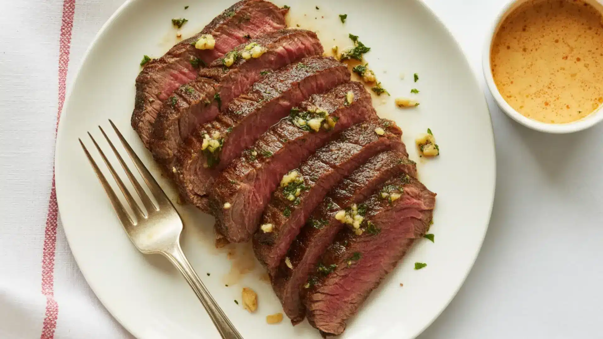 sliced broiled flank steak topped with garlic and herbs on a white plate with a side of creamy sauce