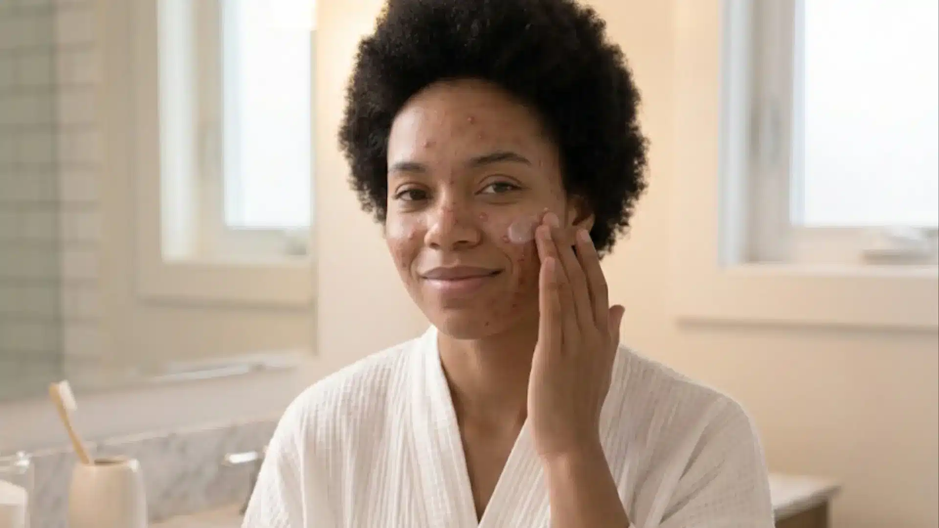 smiling woman with acne prone skin applying cerave moisturizer to her cheek in a bright white bathroom