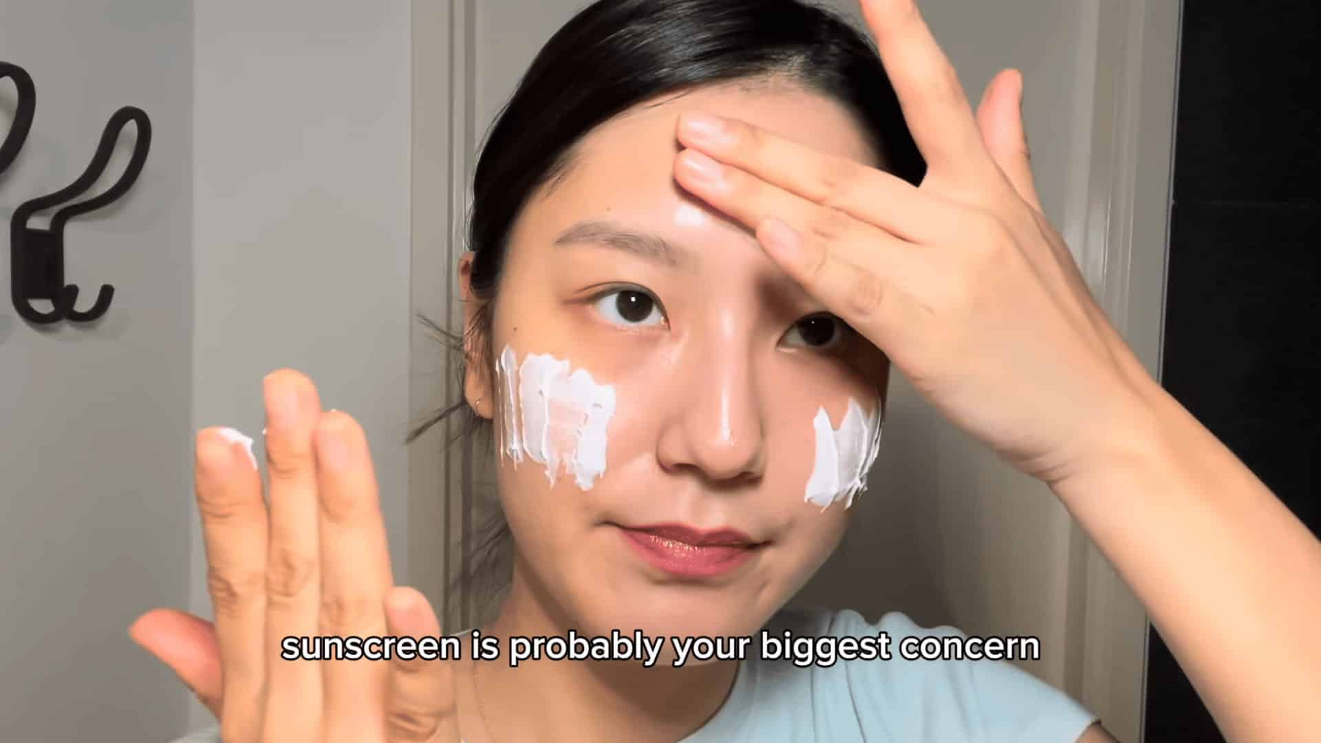 woman applying sunscreen to forehead and cheeks with both hands in bathroom with towel rack in background