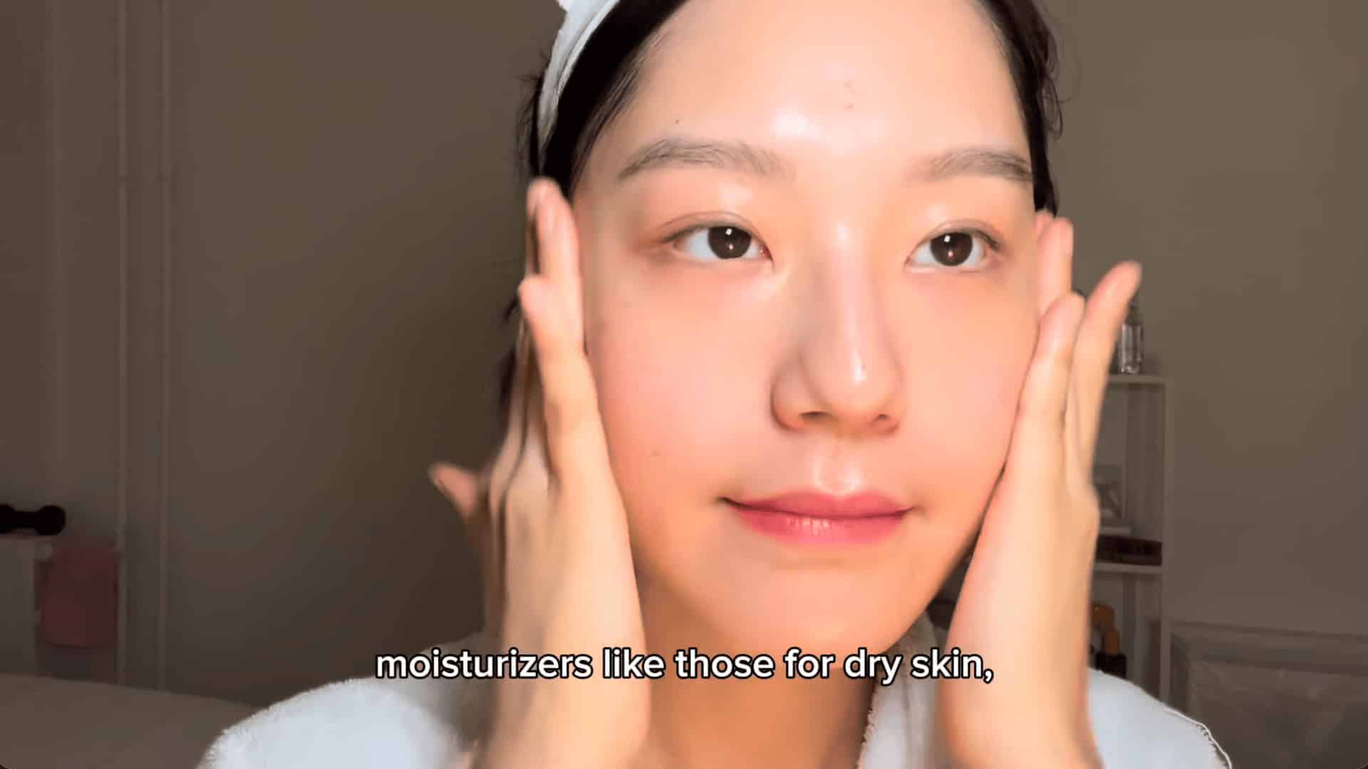 woman gently pressing moisturizer into her cheeks with both hands in a softly lit bedroom with skincare shelf behind her