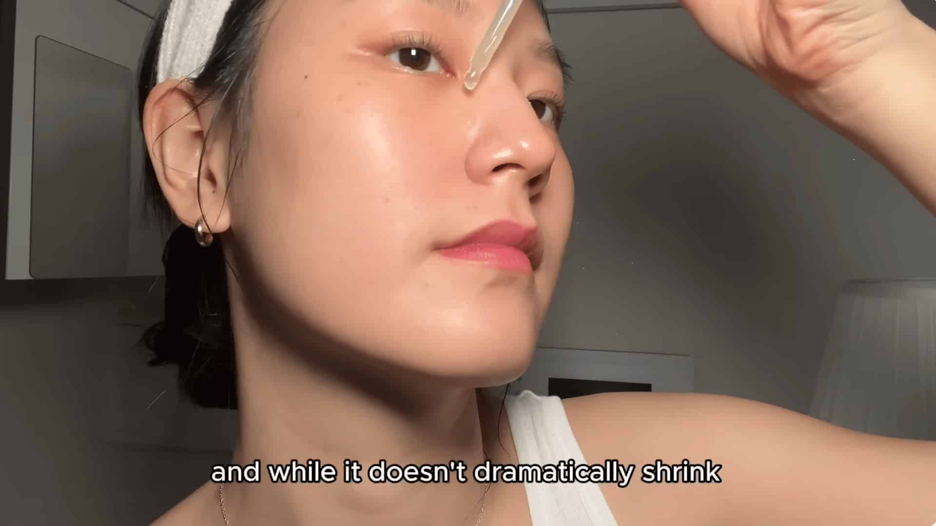 young woman applying facial serum drop to forehead with dropper indoors wearing white headband close up side view