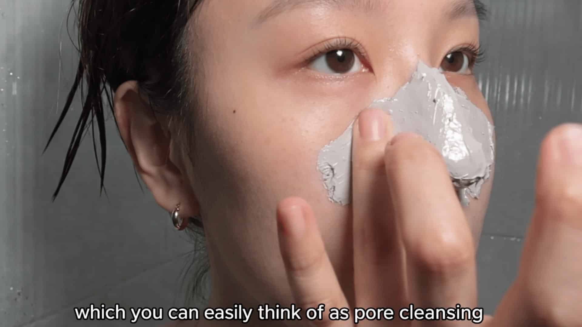 young woman applying gray clay mask to her nose in bathroom close up with wet hair and tiled wall background