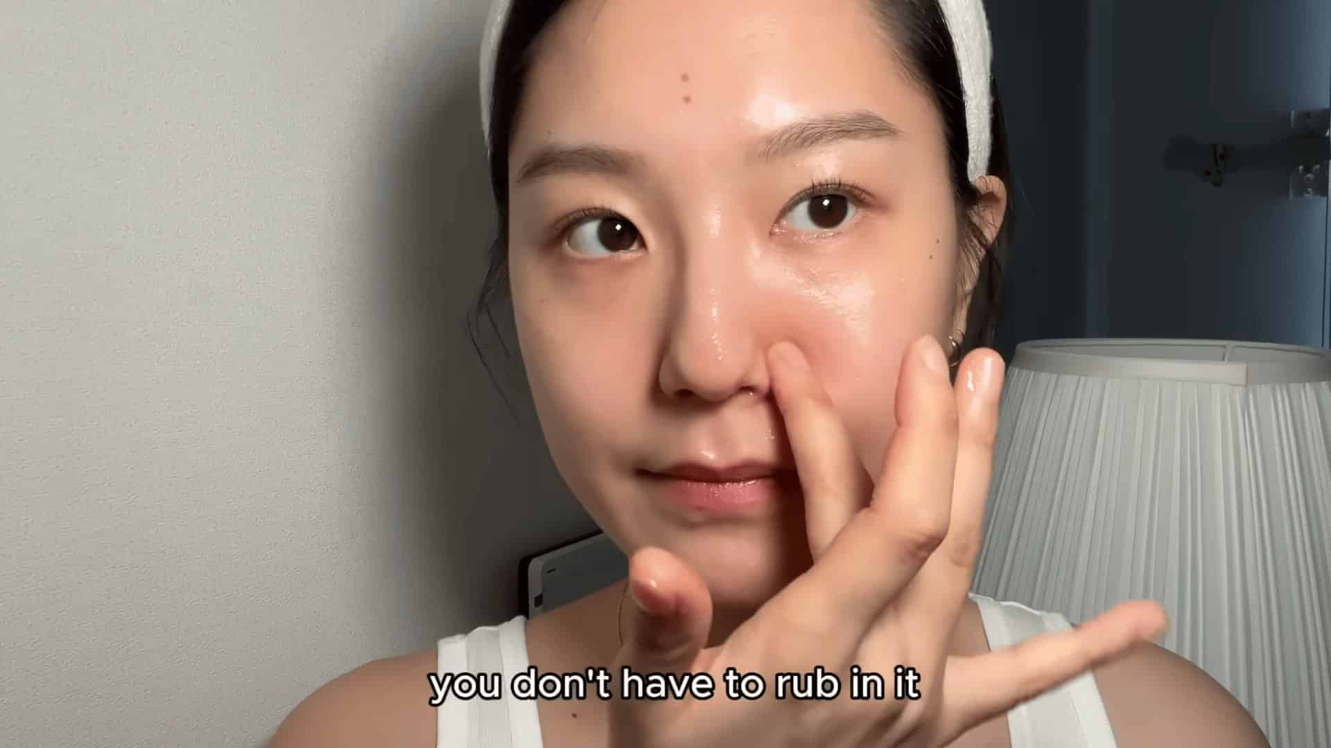 young woman applying skincare serum to her nose indoors with soft lighting and lamp in background close up