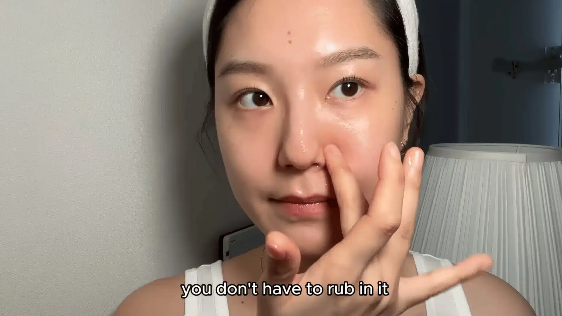 young woman applying skincare serum to her nose indoors with soft lighting and lamp in background close up