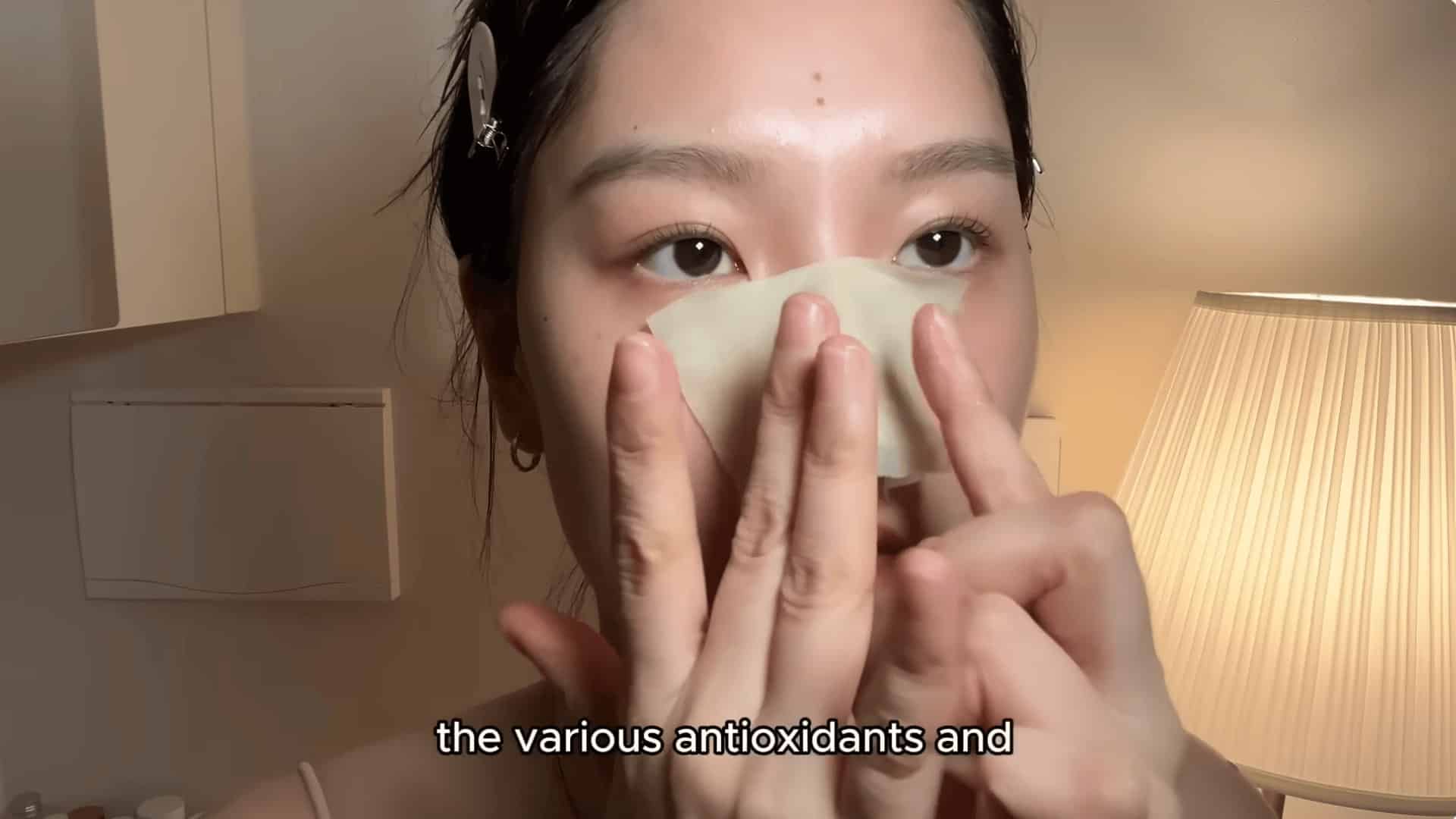 young woman pressing pore strip onto her nose indoors with soft lamp lighting and hair clips visible close up