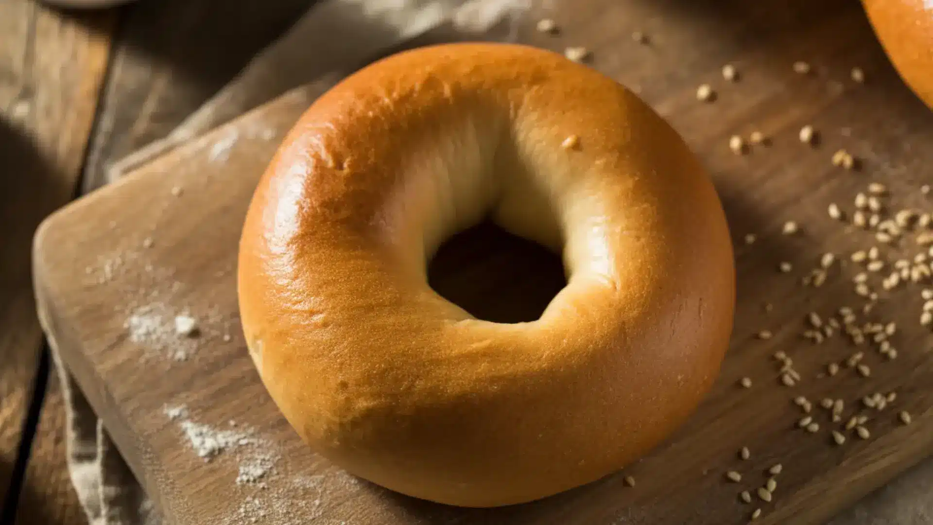 a single golden brown plain bagel sitting on a wooden cutting board with sesame seeds scattered in the background