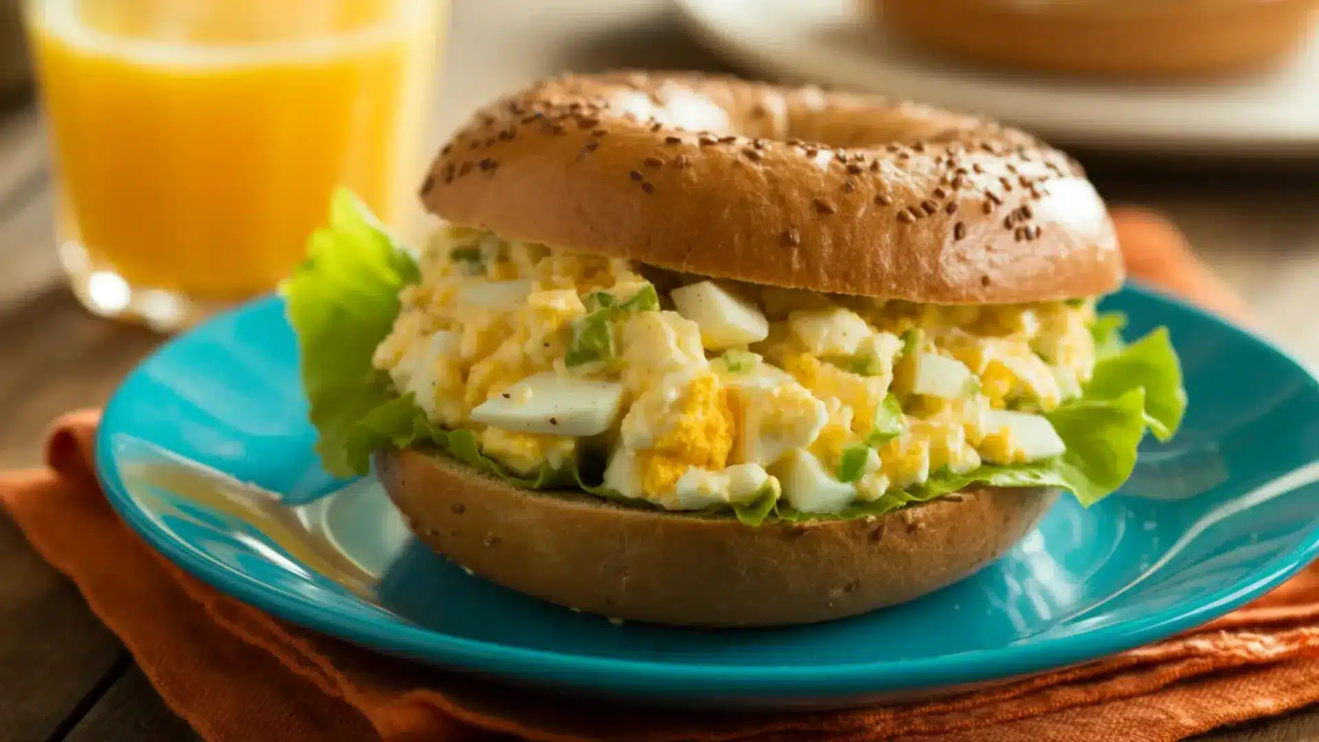 egg bagel sandwich filled with creamy egg salad and green lettuce on a blue plate with orange juice in the background