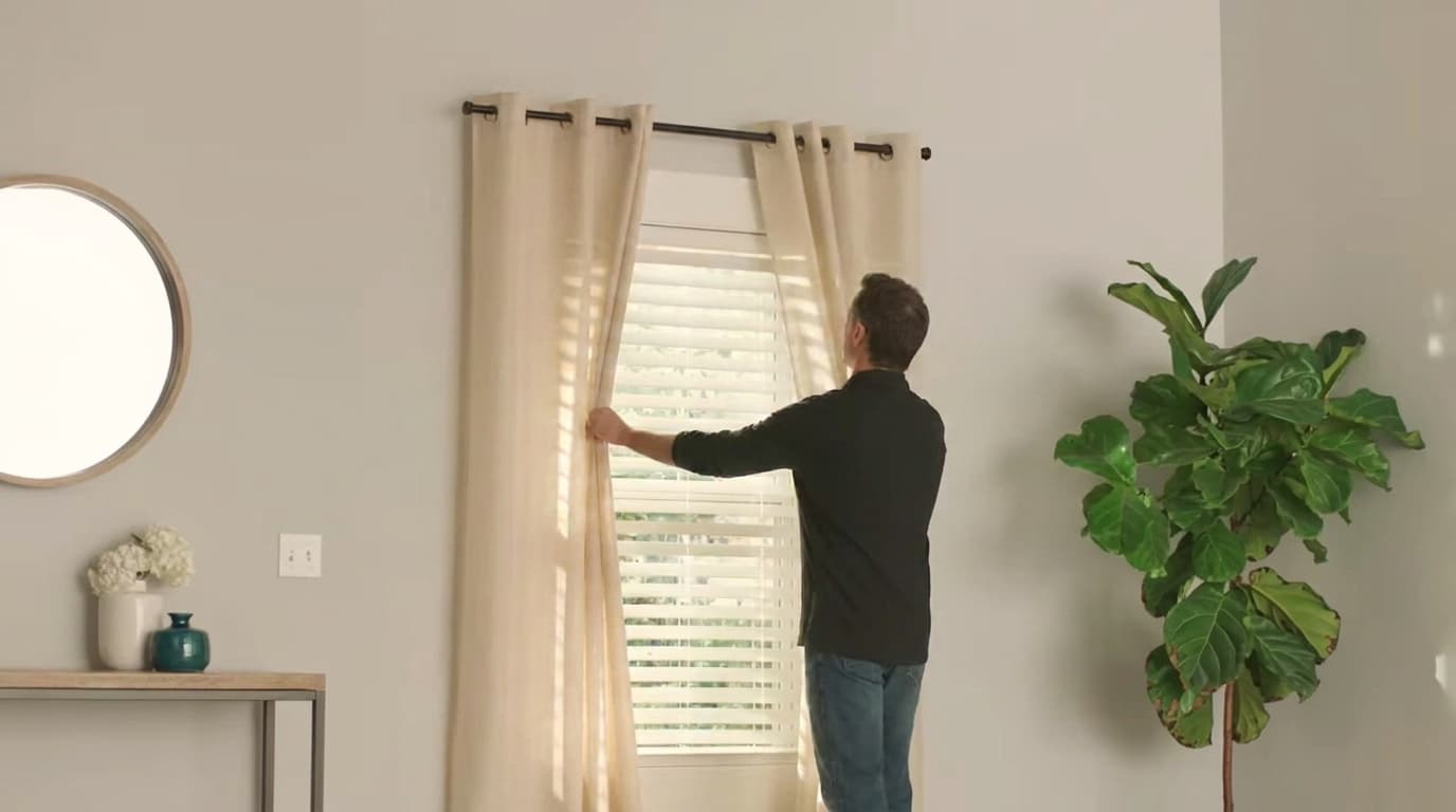 man adjusting cream grommet curtains hanging over white blinds on a black curtain rod in a bright living room