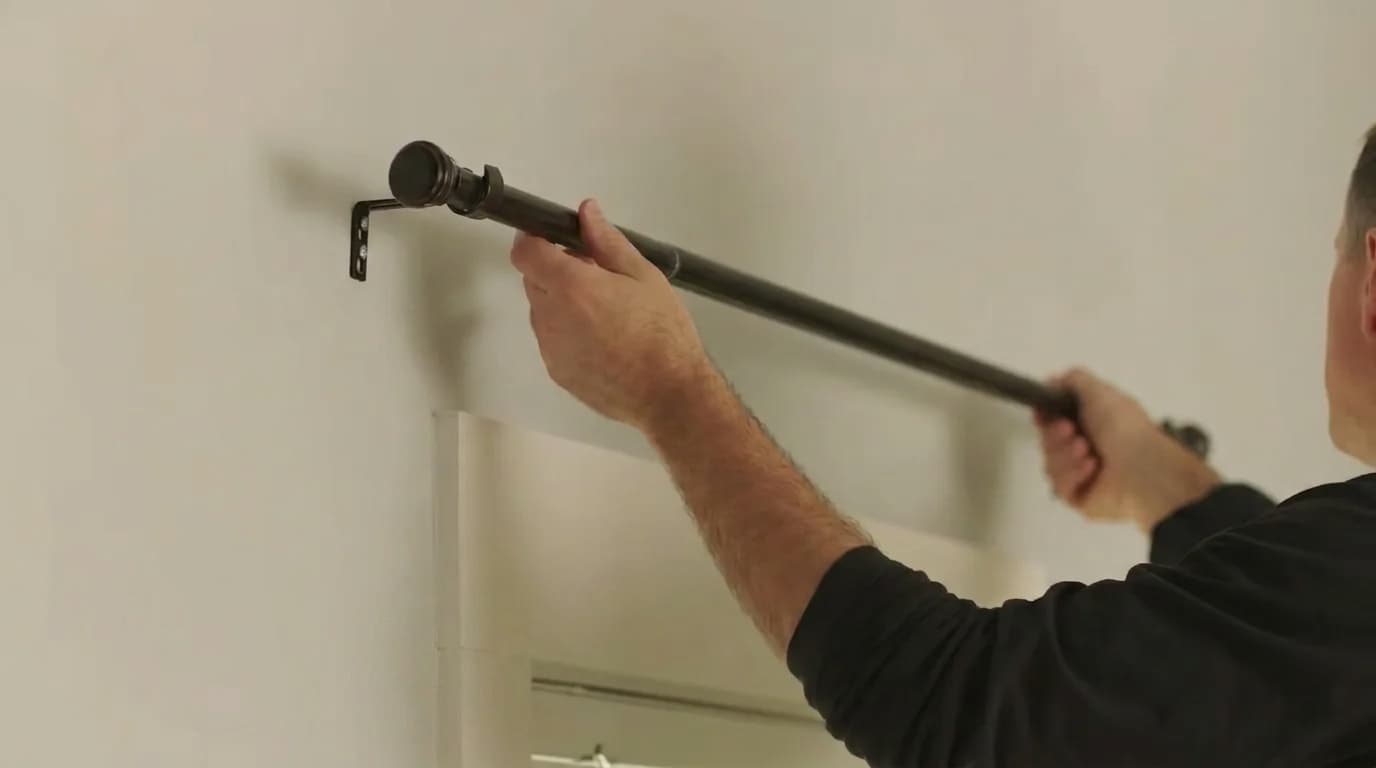 man placing a dark curtain rod onto a wall mounted bracket above a window during curtain installation