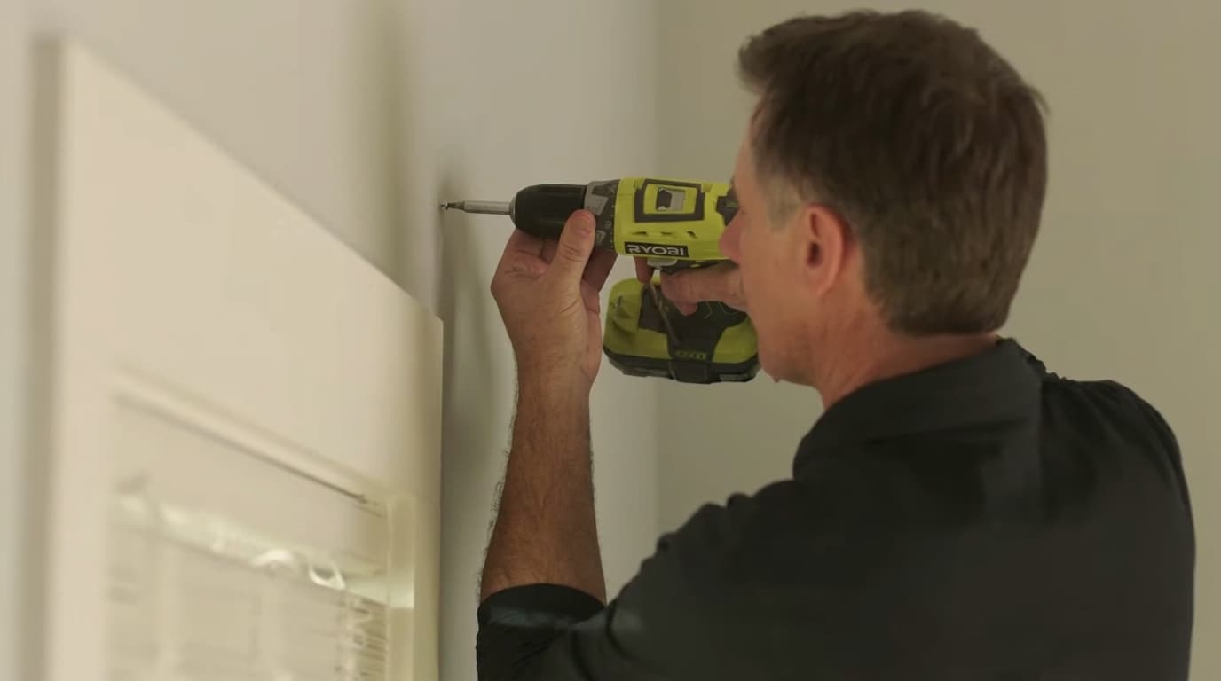 man using a ryobi cordless drill to drill guide holes into a wall beside a window frame for curtain bracket installation