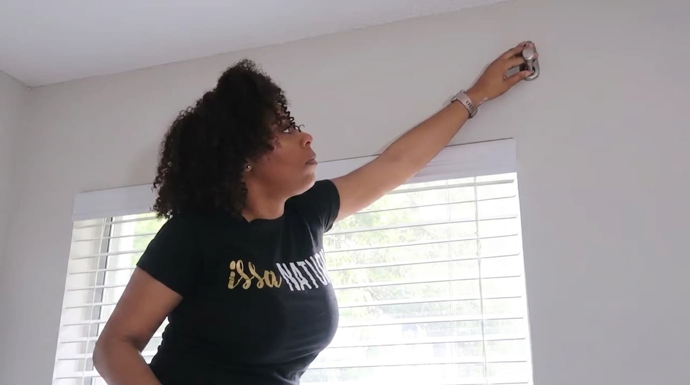 woman reaching up to mounting the command hooks on ceiling inside room near window blinds