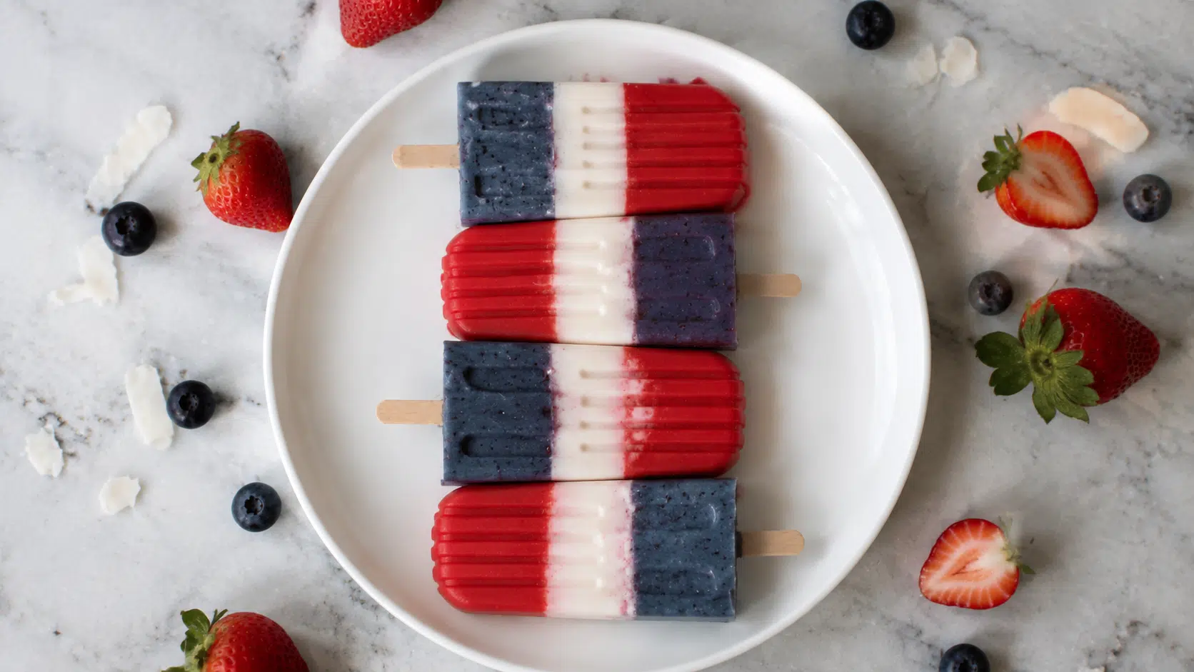 Four red, white, and blue popsicles arranged on a white plate, surrounded by fresh strawberries and blueberries on a light marble surface