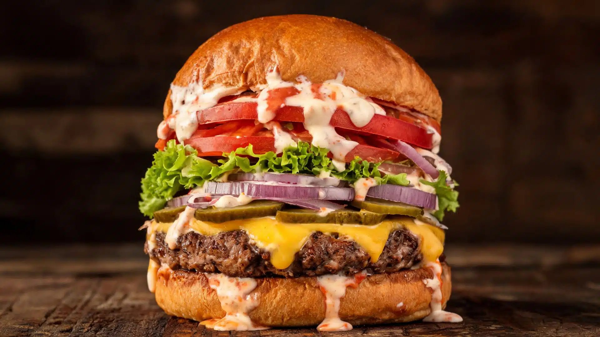 loaded gourmet burger stacked with cheese lettuce tomato onion pickles and creamy sauce dripping on toasted bun rustic background
