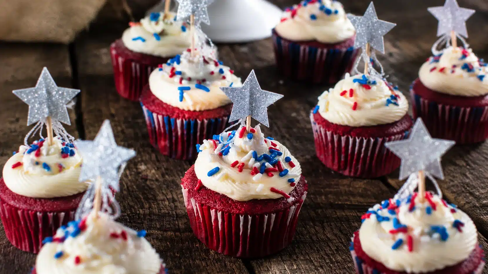 Red velvet cupcakes topped with white frosting, red blue sprinkles, and silver star decorations on a wooden surface